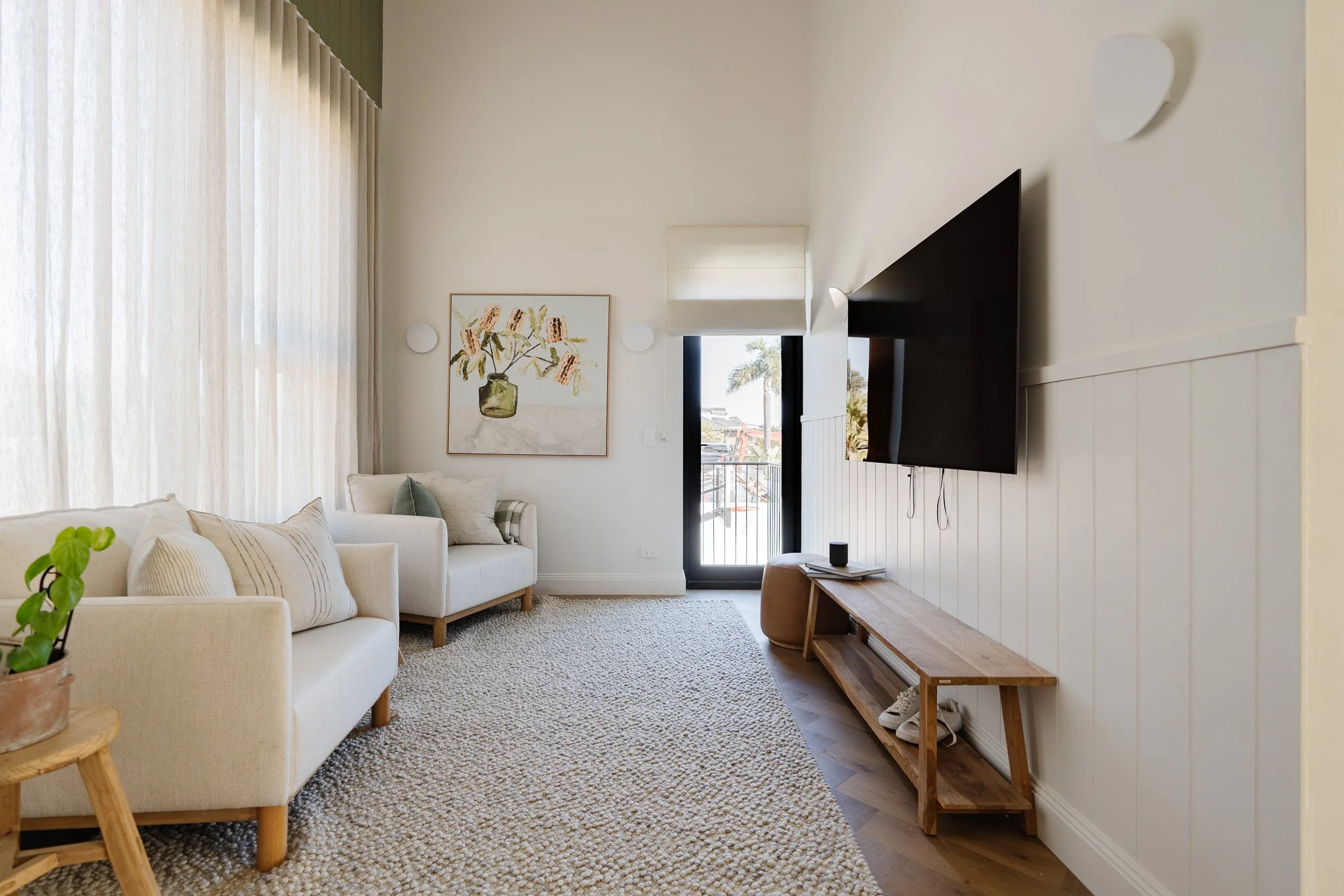 Living room with two white sofas, a textured rug, a wall-mounted TV, a small wooden console, a sliding glass door, and a framed artwork of a vase with flowers on the wall.