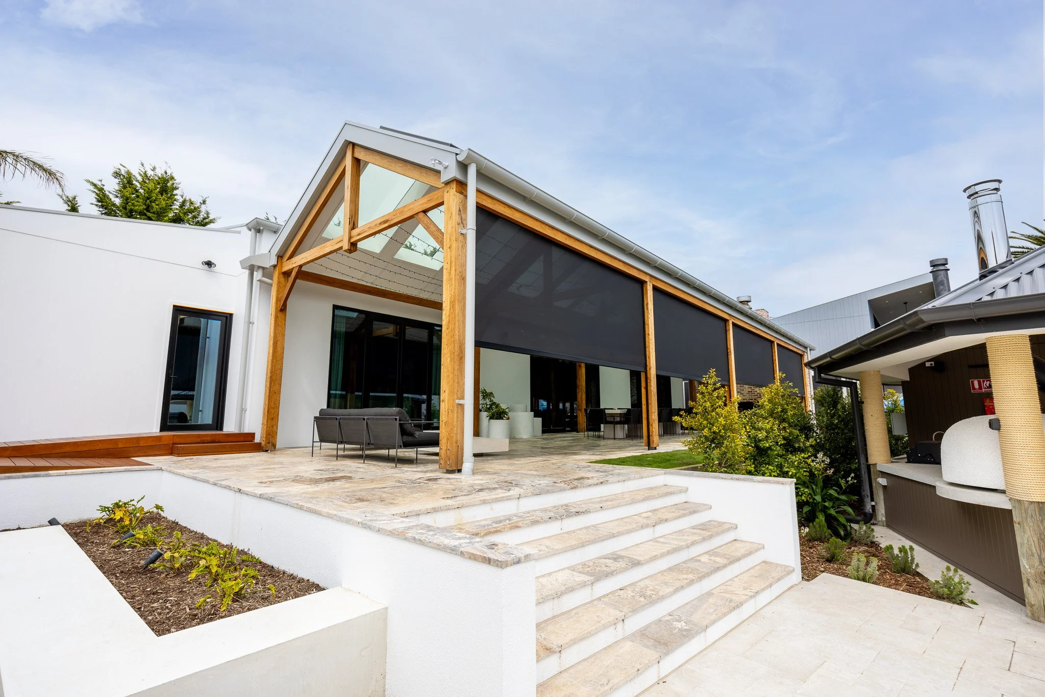 Modern house with white exterior, wooden beams, large glass windows, and a patio with outdoor furniture, stairs, and landscaping.