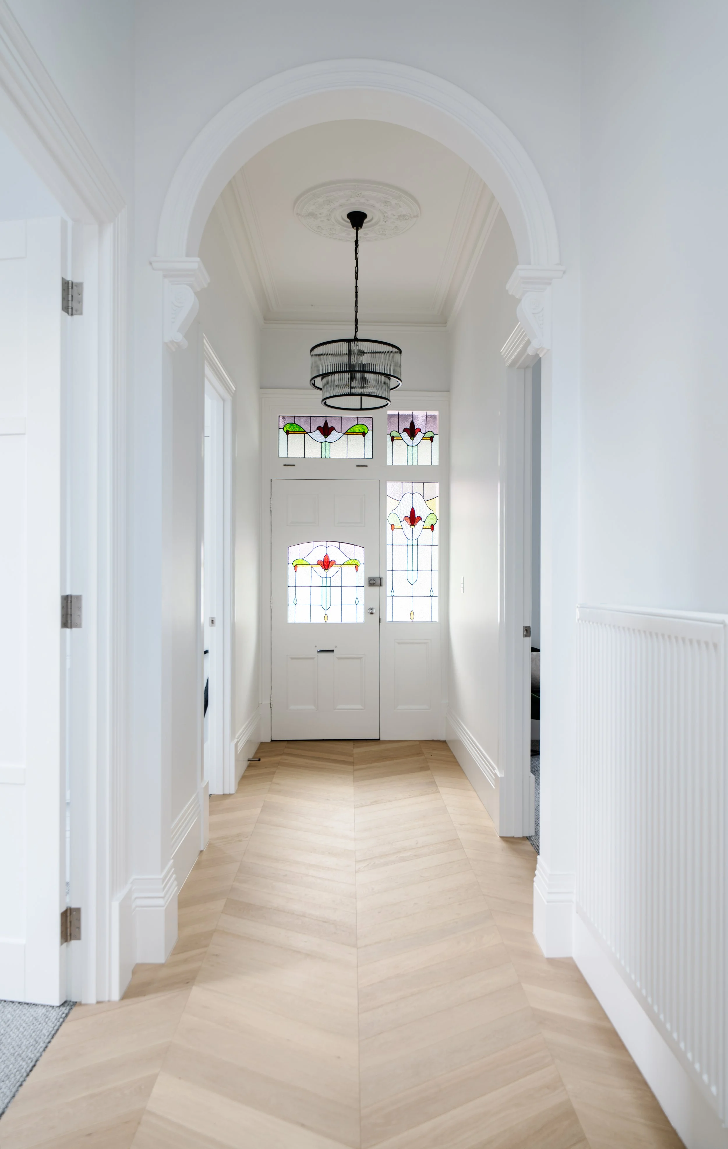 Bright, elegant entryway with a white door featuring stained glass panels, a modern chandelier hanging from a decorative ceiling medallion, and light wooden chevron-patterned flooring.