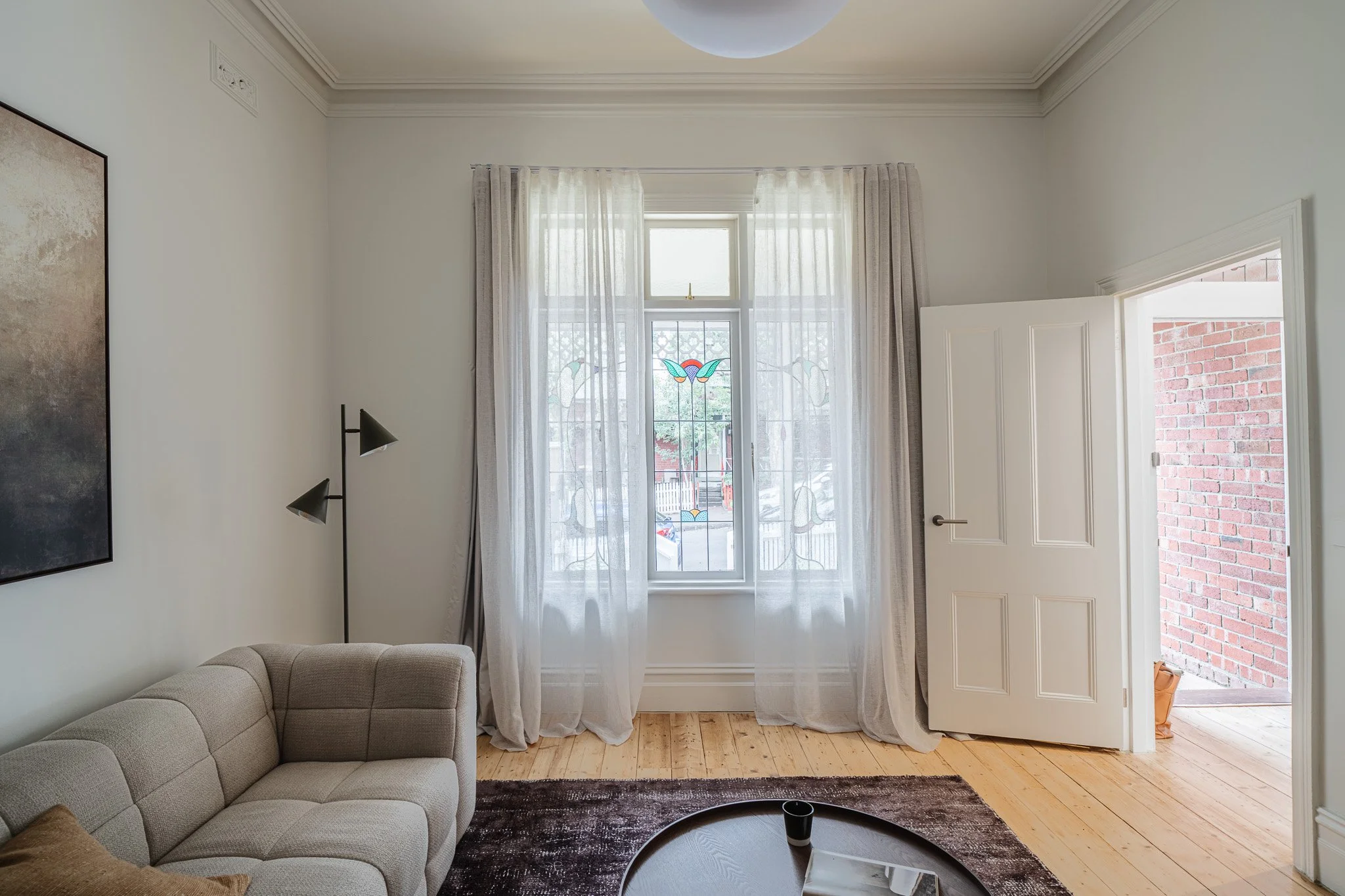 Living room with beige sofa, floor lamp, window with sheer curtains, open door to the outside brick wall, and a small round coffee table.