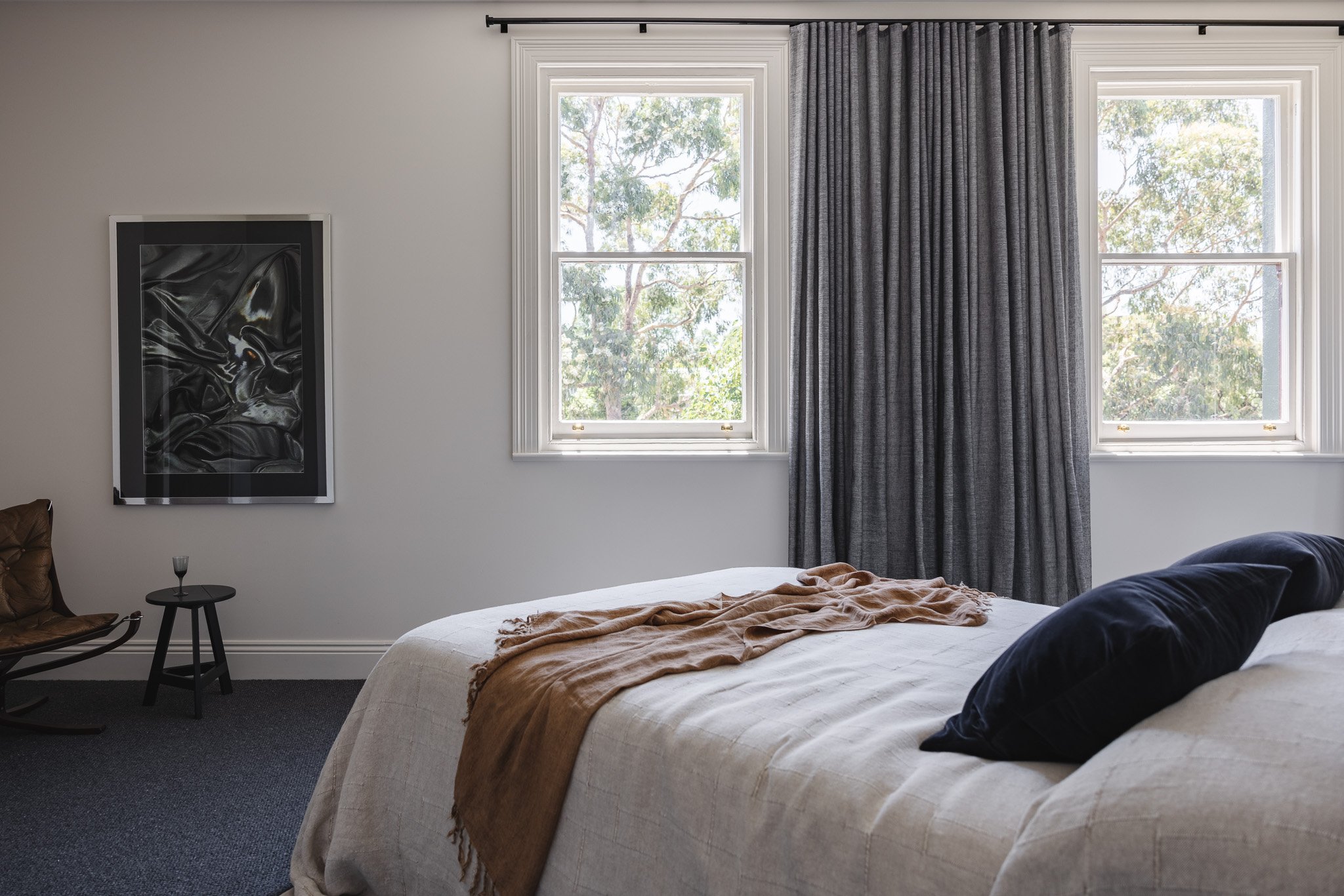 A bedroom with two large windows, framed by white trim and gray curtains, a bed with beige bedding and dark pillows, a small black side table, a brown leather chair, and a black abstract artwork on the wall.