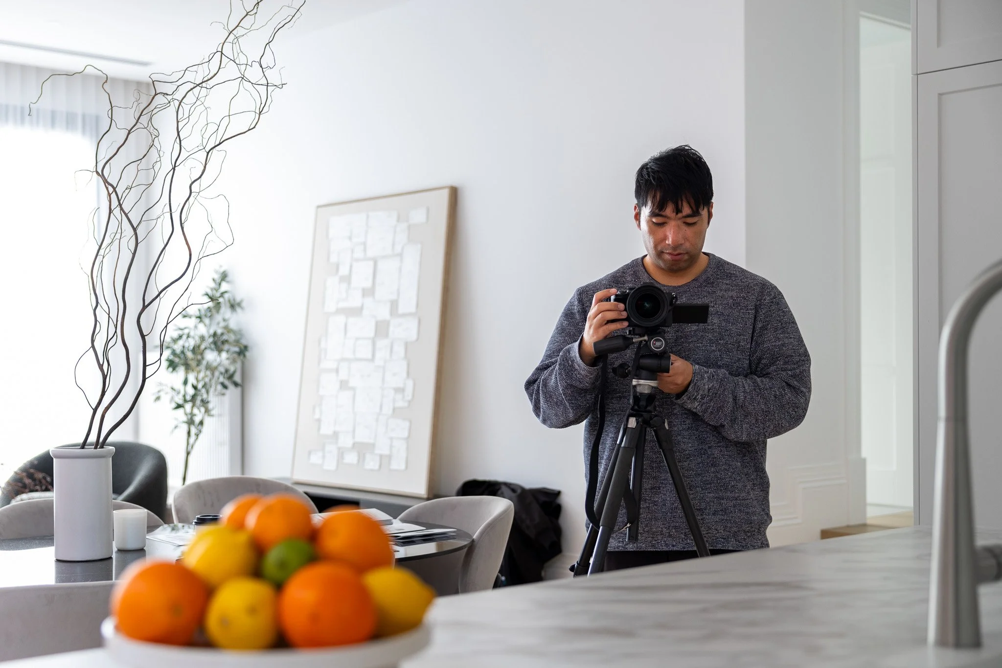 A man takes a photograph with a camera on a tripod in a modern, well-lit home kitchen or dining area, with a bowl of oranges and lemons on the counter in the foreground and minimalist decor in the background.
