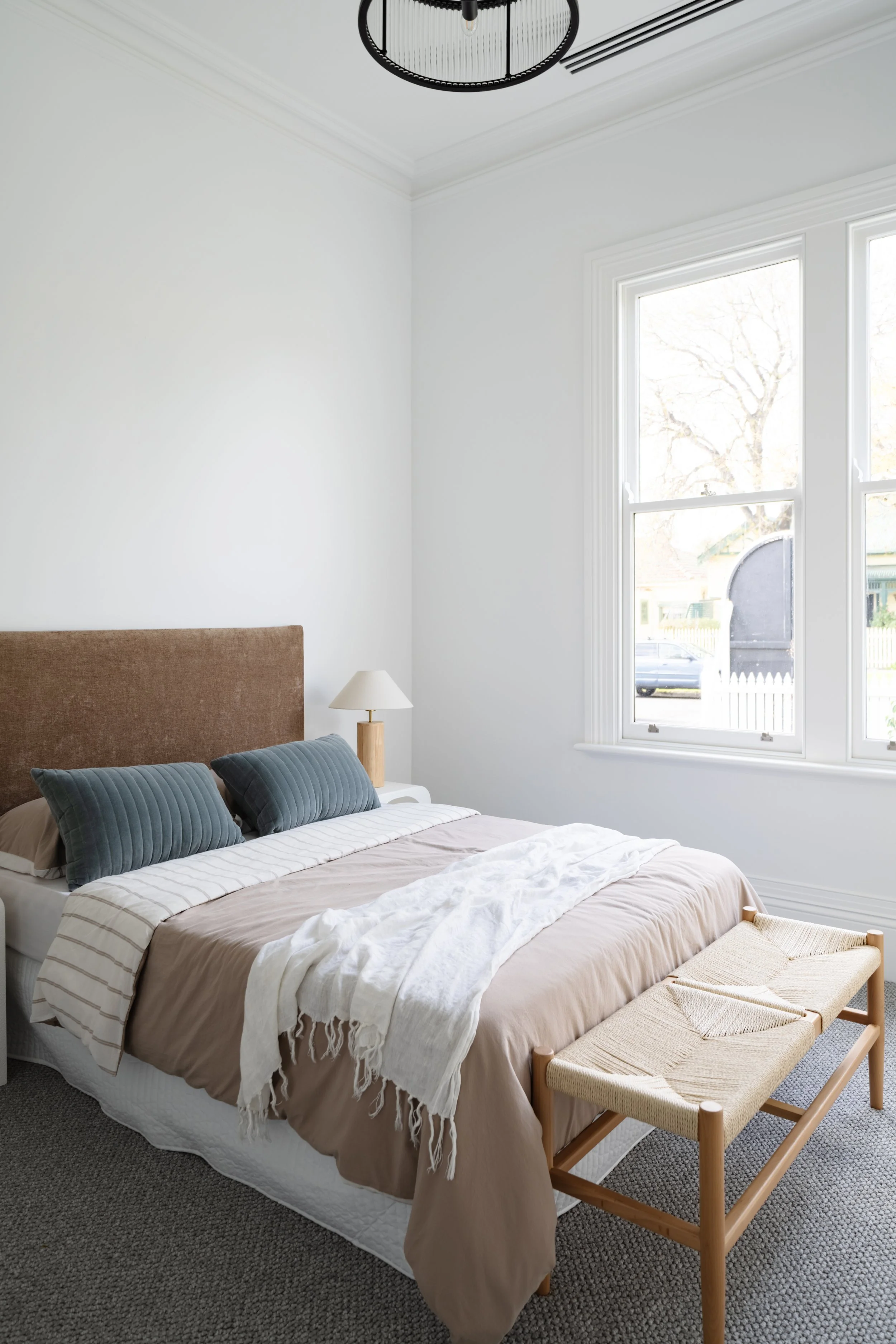 A neatly made bedroom with a beige bed, two dark teal pillows, a white throw blanket, a small white bedside table with a lamp, a woven bench at the foot of the bed, and large windows with white trim letting in natural light.