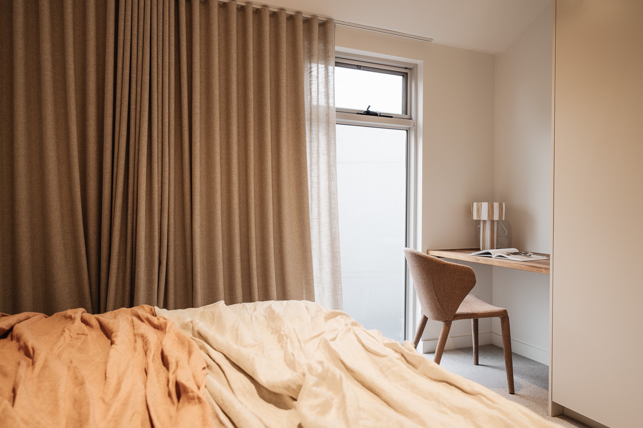 A cozy bedroom corner featuring beige curtains, a window, a small wooden desk with a striped lamp, an open book, and a brown upholstered chair. The bed has rumpled light-colored sheets.