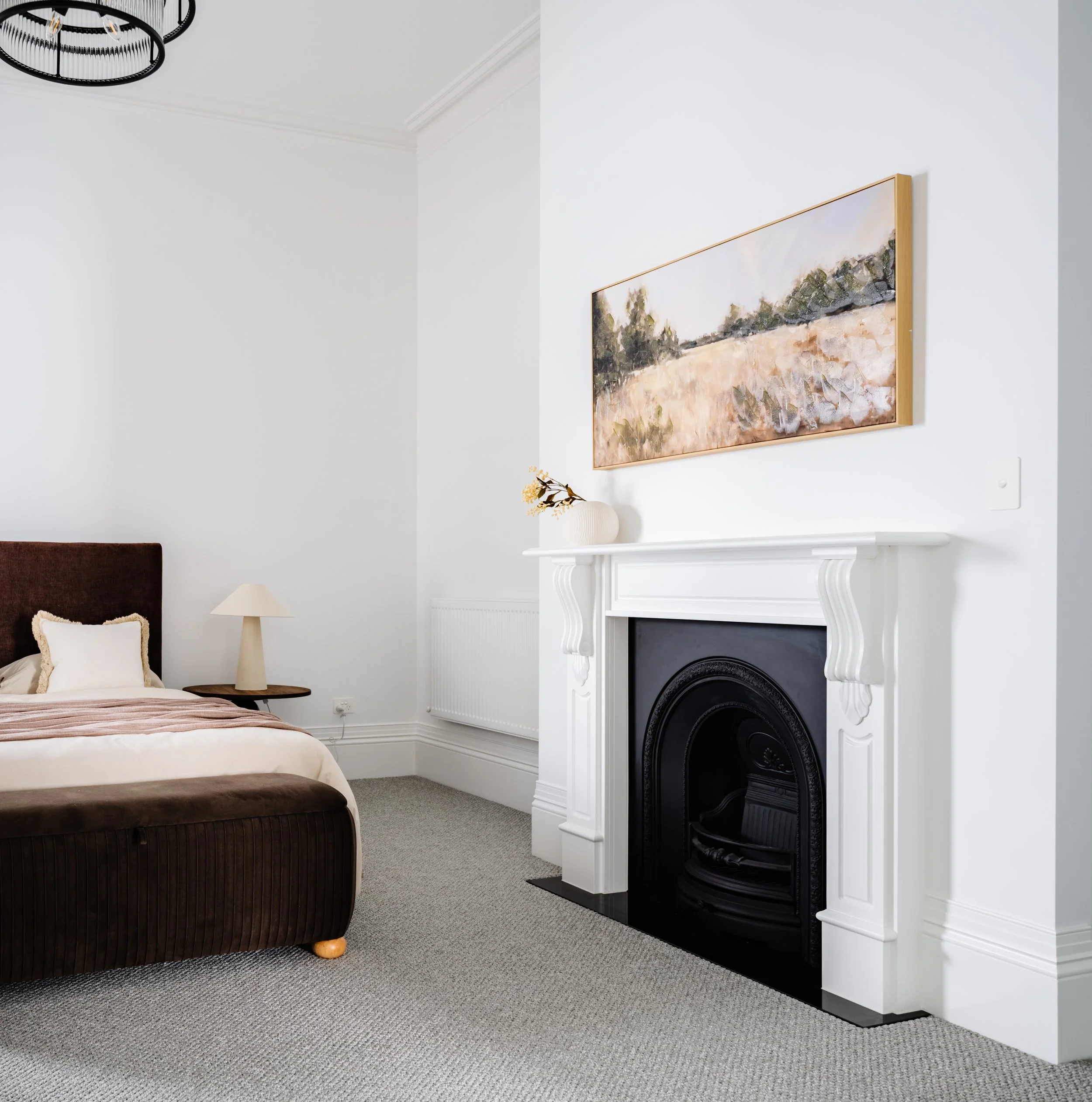 A bedroom with a bed, bedside table, lamp, a decorative vase with flowers, a white fireplace with black insert, a landscape painting above the fireplace, and light-colored walls and carpet.