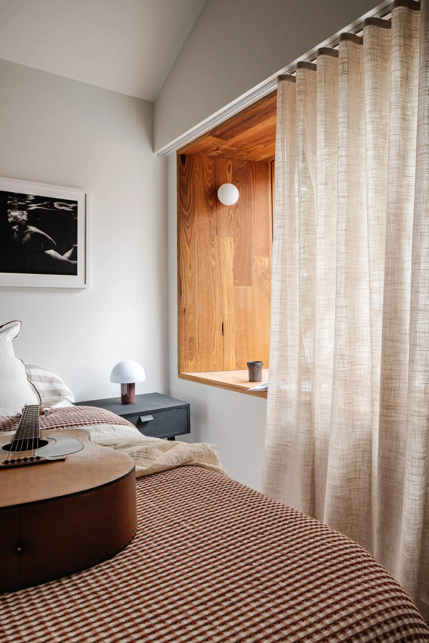 A bedroom with a guitar on the bed, a lamp on the nightstand, a framed black and white photograph on the wall, and a window with beige curtains.