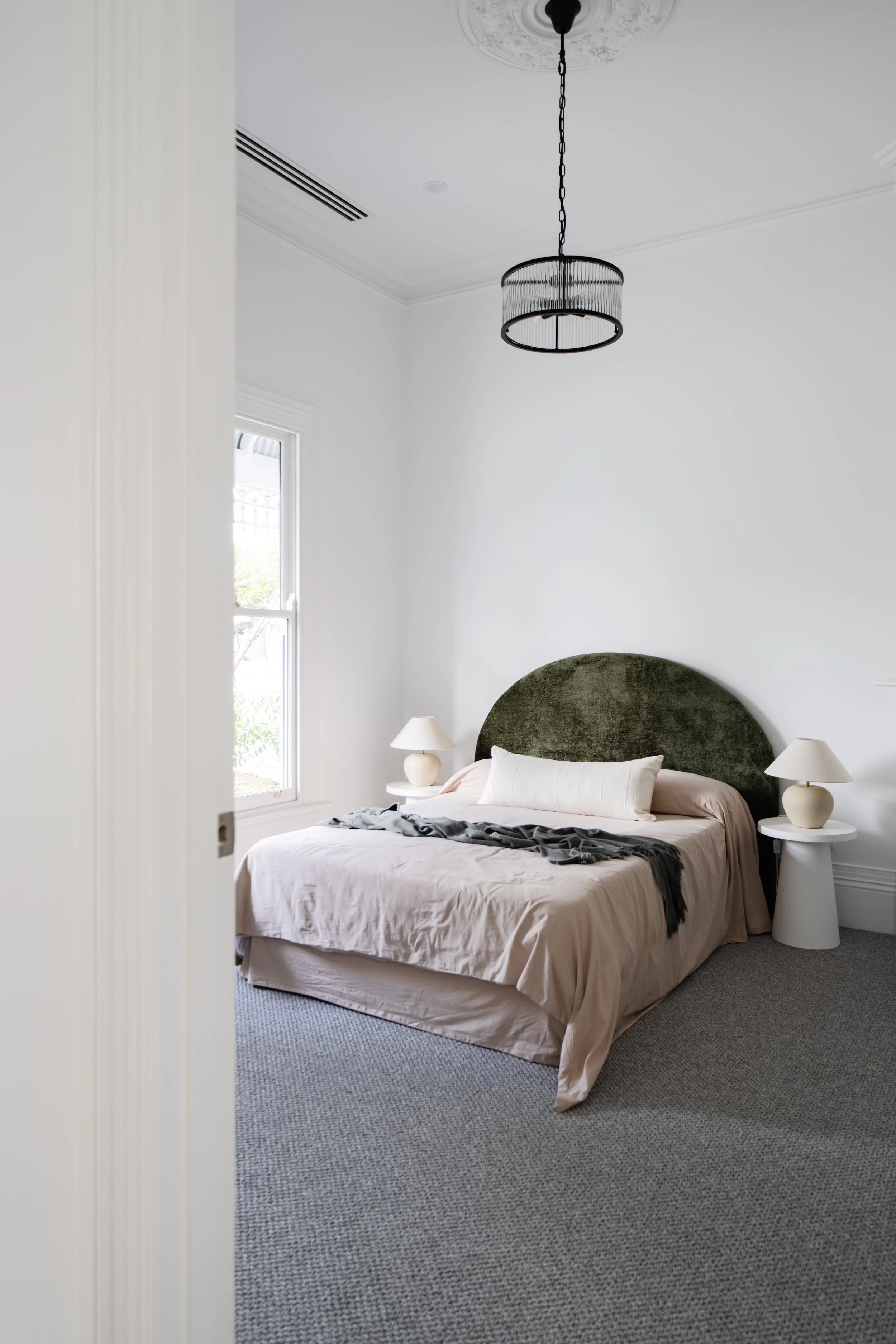 A bedroom with a bed, green velvet headboard, two white nightstands, and matching white lamps, illuminated by natural light through a window.