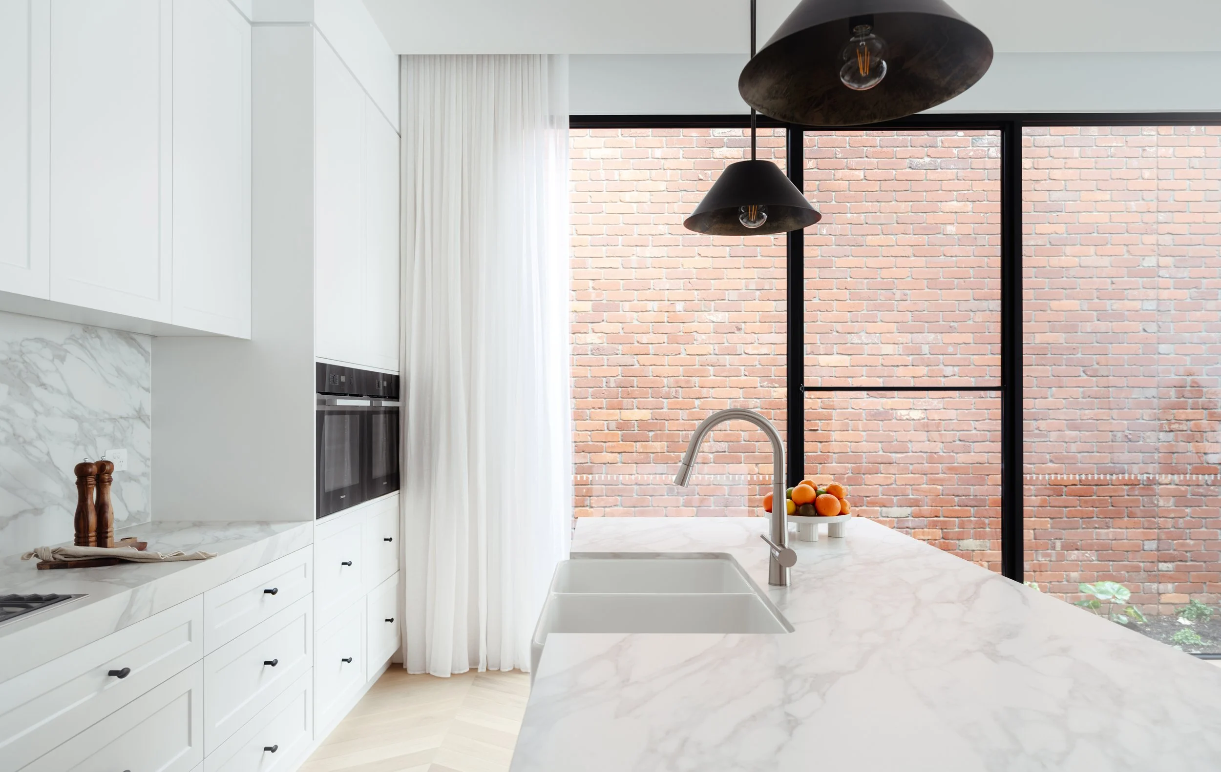 Modern kitchen with white cabinets, marble countertops, and large window with brick wall outside. Black pendant lights and a bowl of fruit on the counter.