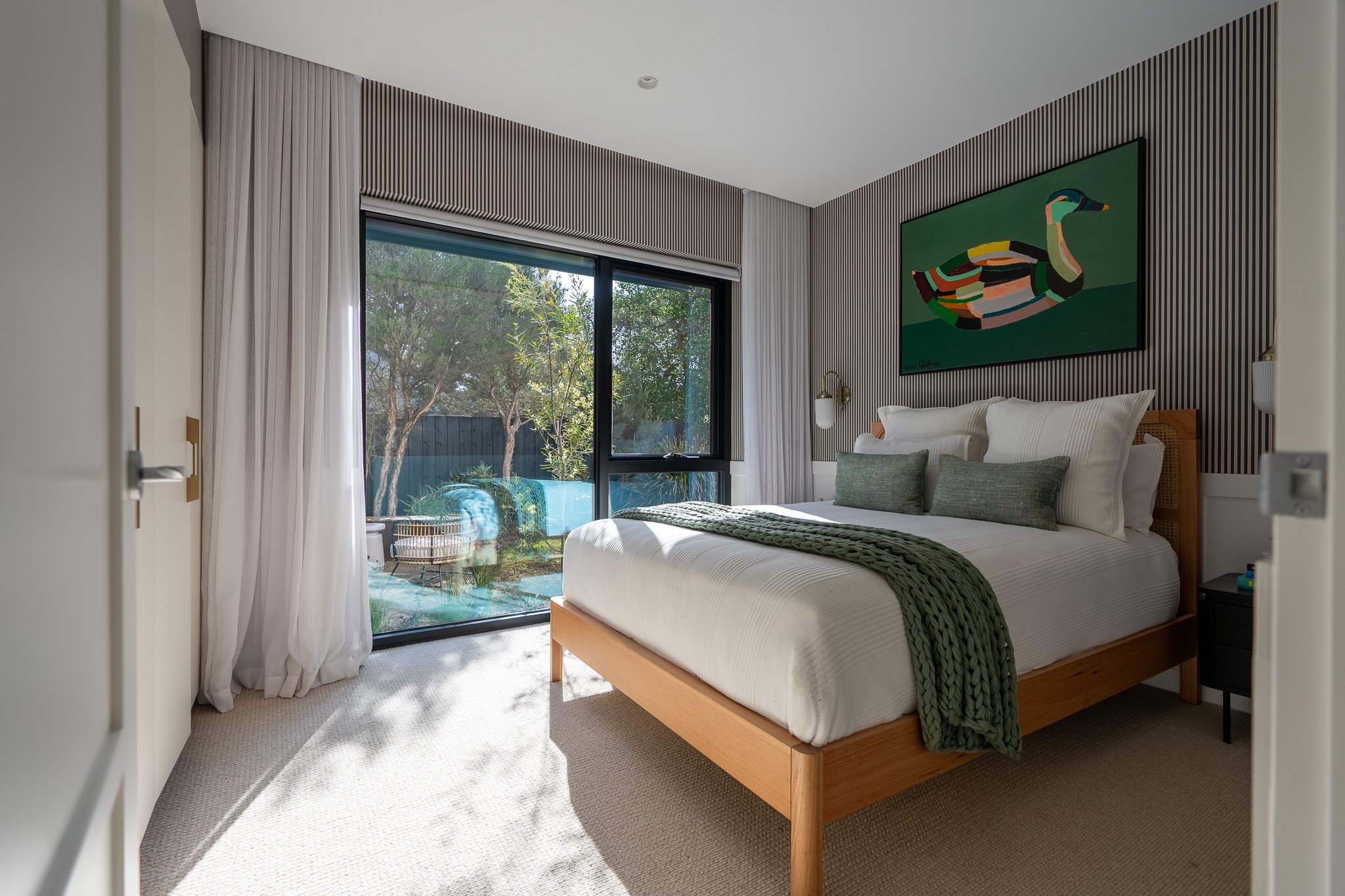 Modern bedroom with a large bed, white curtains, wood accents, and a sliding glass door leading to an outdoor garden.