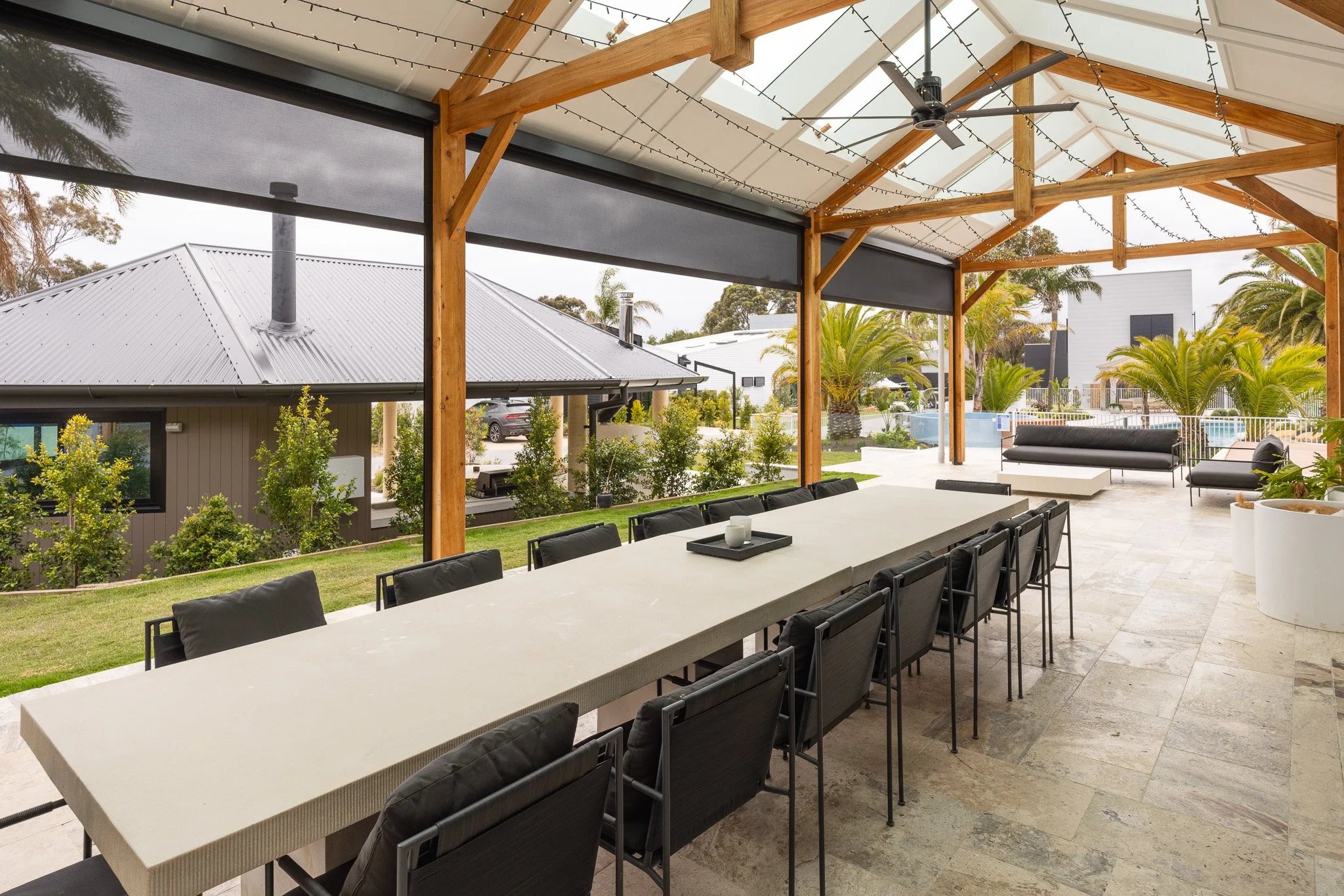 Covered outdoor patio with a long dining table, black chairs, black sofas, and string lights, overlooking a landscaped yard with trees and neighboring house.