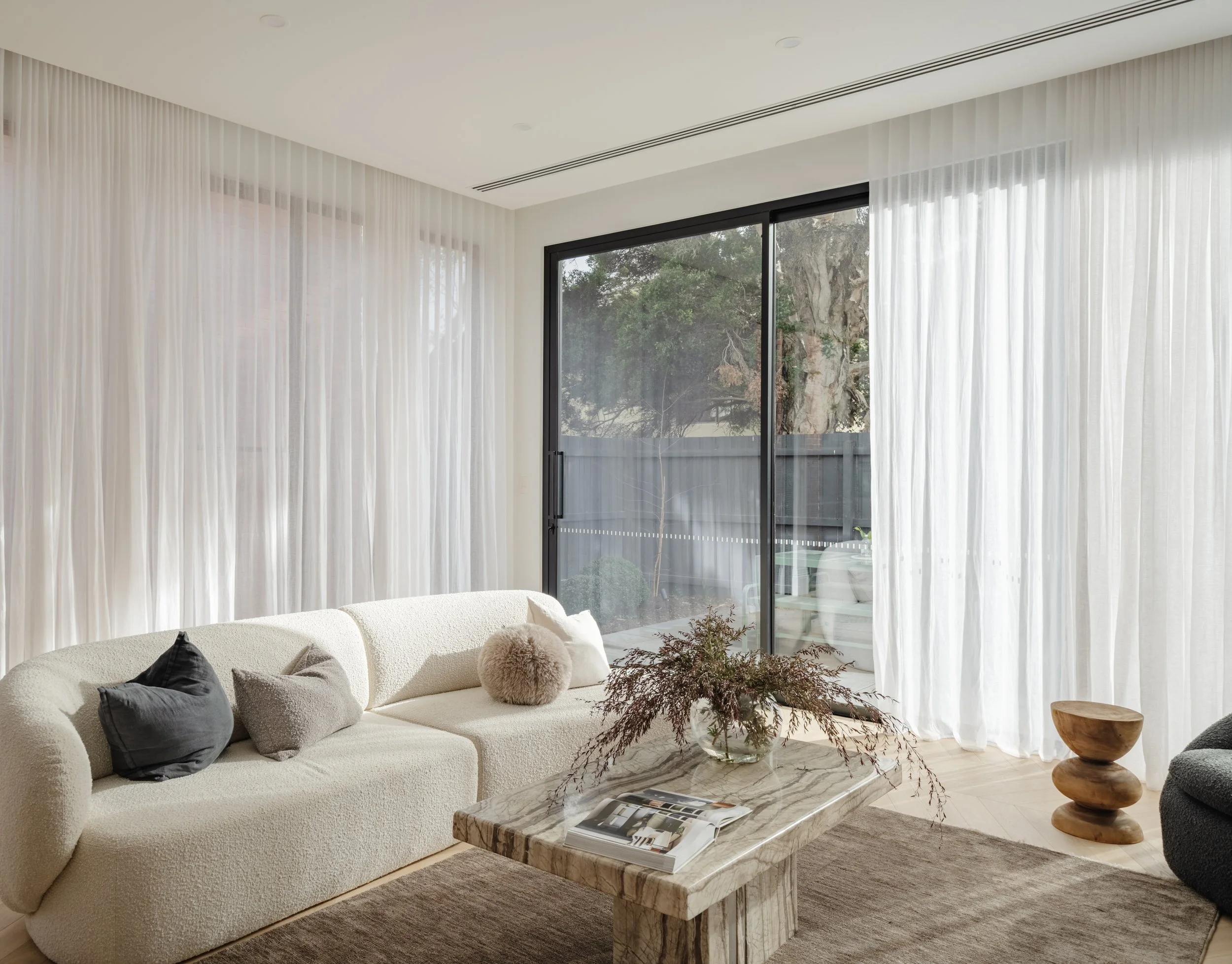 Modern living room with white sofa, black and beige cushions, glass sliding door with sheer white curtains, wooden side table, marble coffee table with a magazine and flowers, and a beige rug.