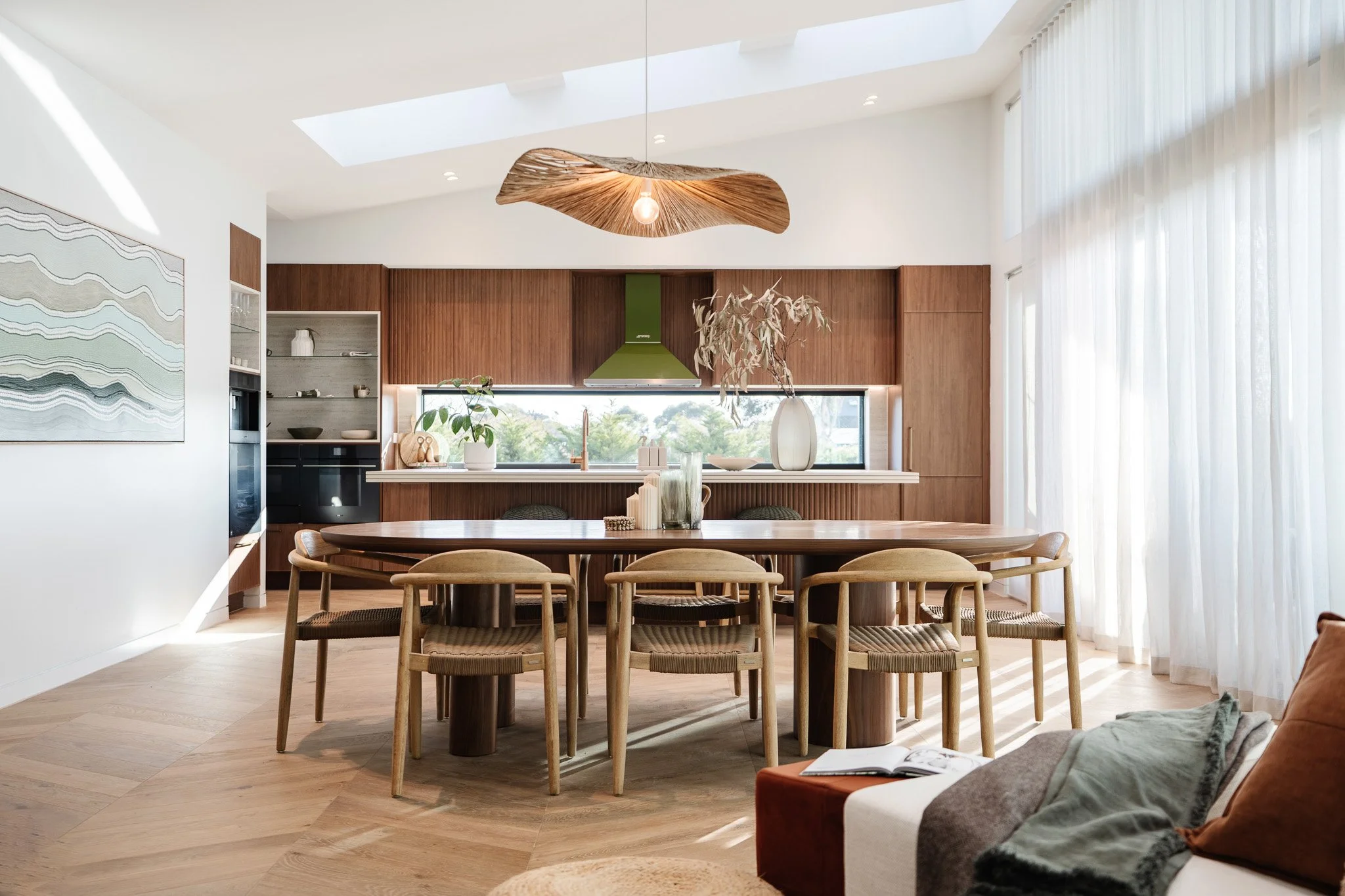 Modern open-concept kitchen and dining room with wooden accents, a large oval dining table with six chairs, a green range hood, a skylight, large windows with sheer curtains, and abstract wall art.