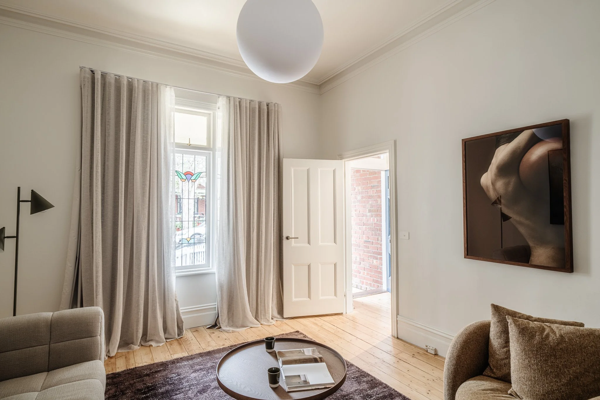 Living room with beige sofas, a wooden coffee table with magazines and cups, a window with sheer curtains, a partially open door, built-in wall art featuring a profile of a woman's face, and a large round ceiling light.