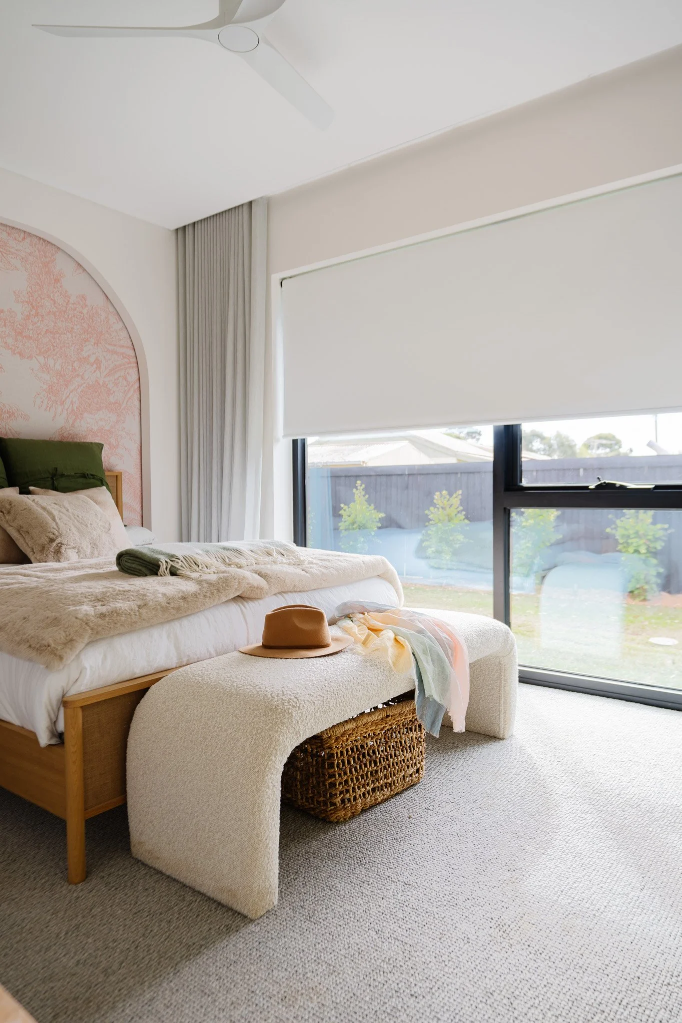 A bedroom with a bed that has pillows, a blanket, and a hat on it, next to a wicker basket and a window with white blinds and light-colored curtains.