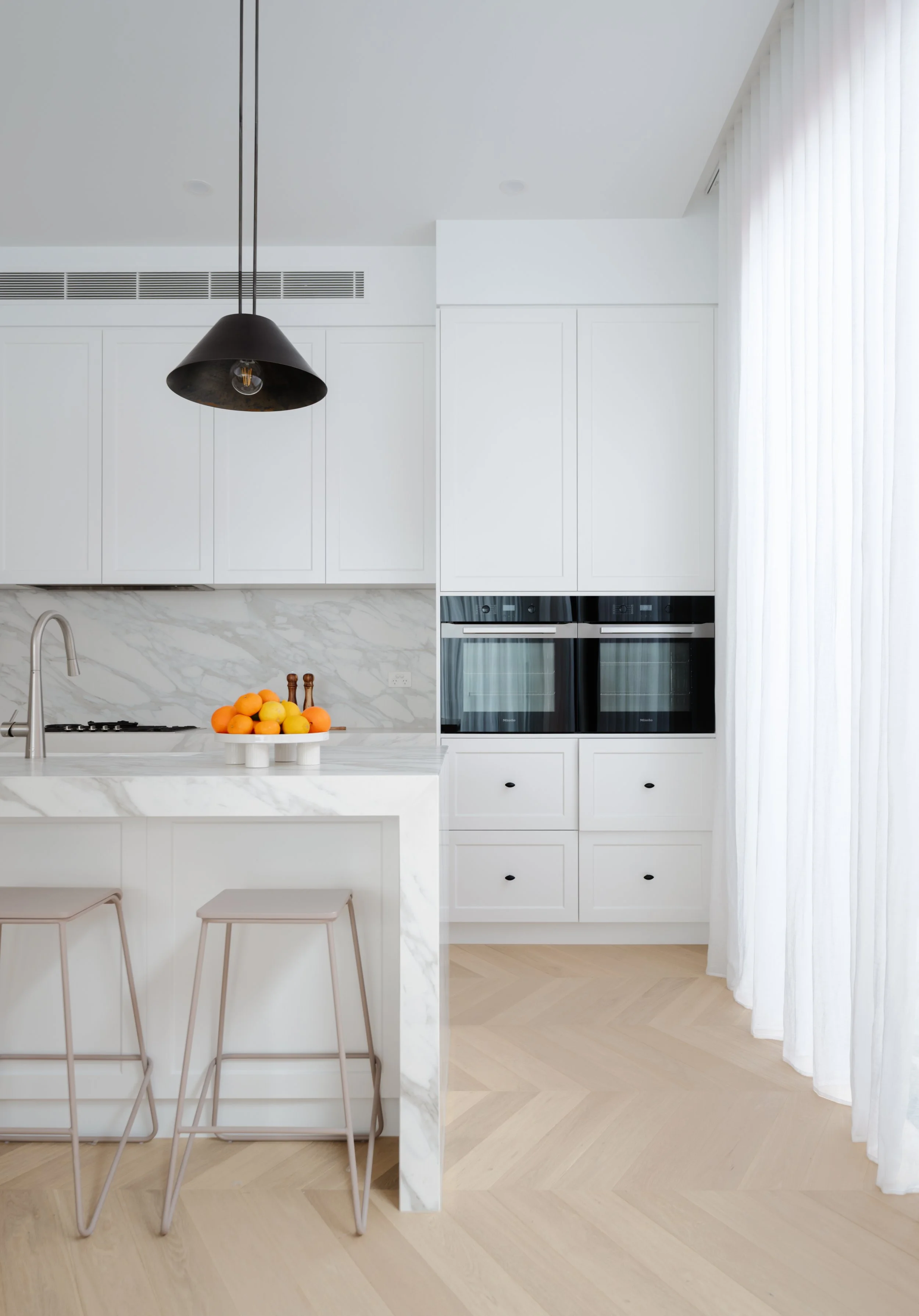 Modern kitchen with white cabinets, marble countertops, a black pendant light, a fruit bowl with oranges and lemons, and a large window with sheer white curtains.