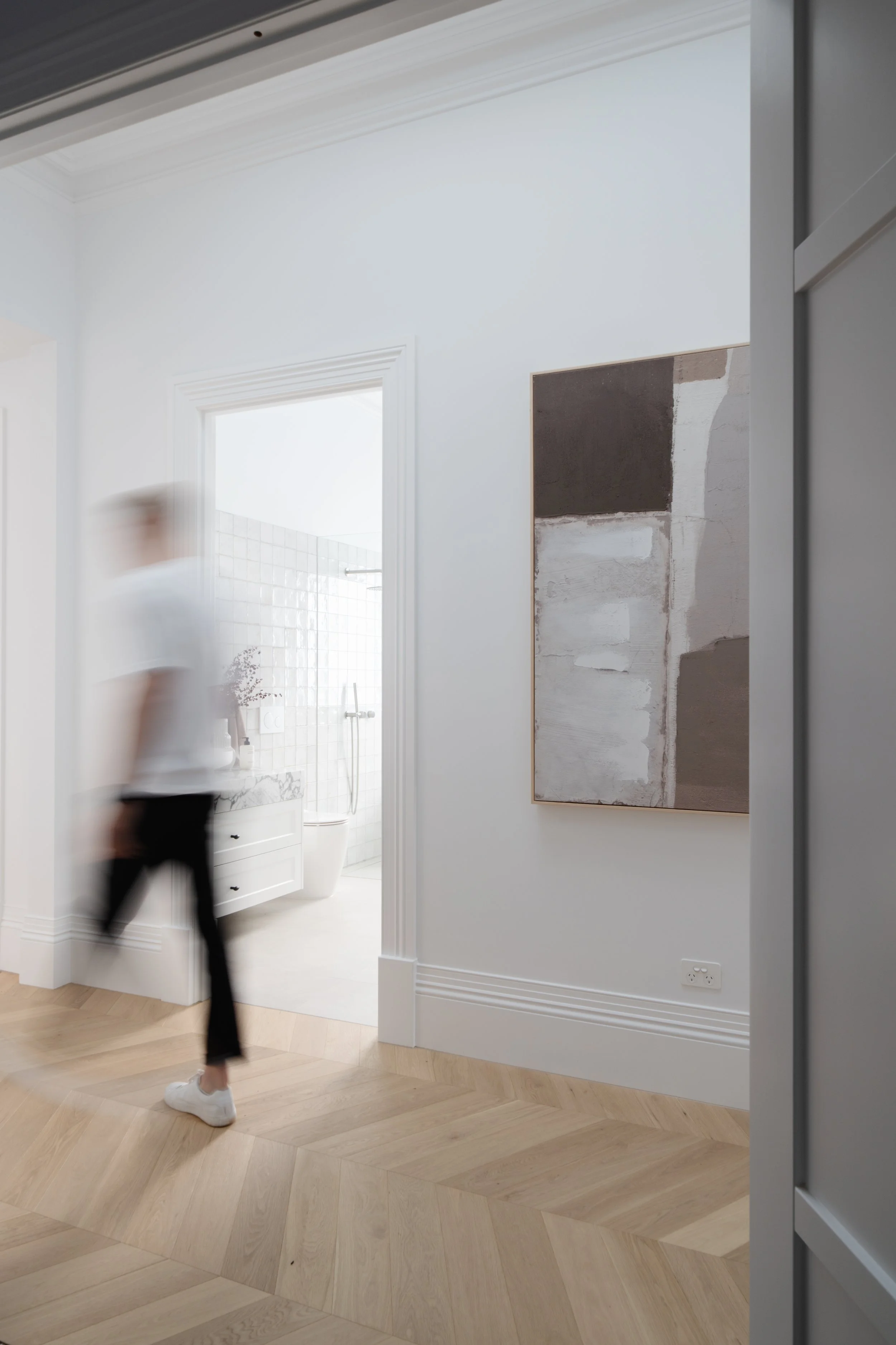 A person walking past a doorway leading to a bathroom, with a modern, abstract painting on the wall and light-colored wooden flooring.