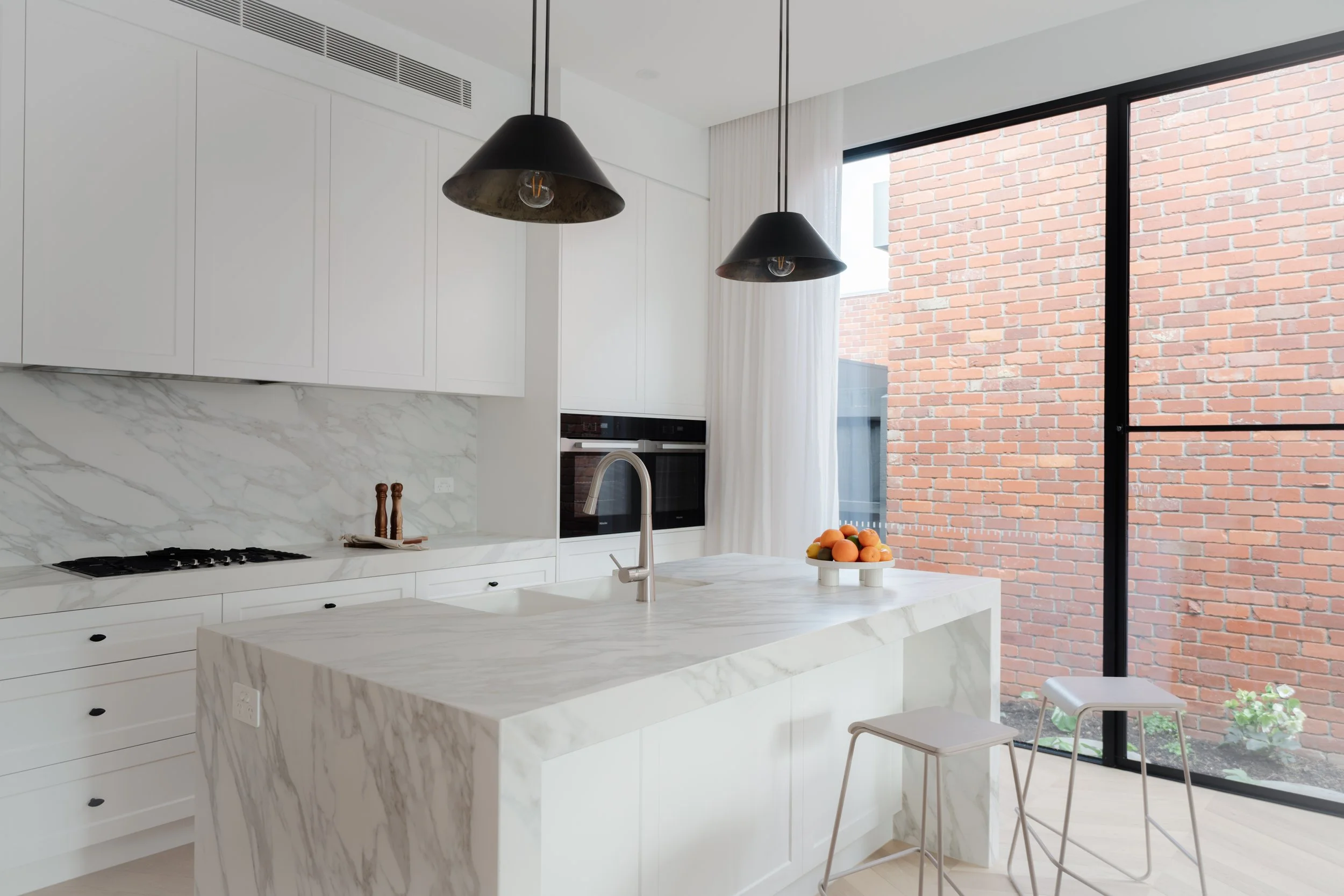 Modern kitchen with white marble island, white cabinets, black pendant lights, and a large sliding glass door showing a brick wall outside, with a bowl of oranges on the island.