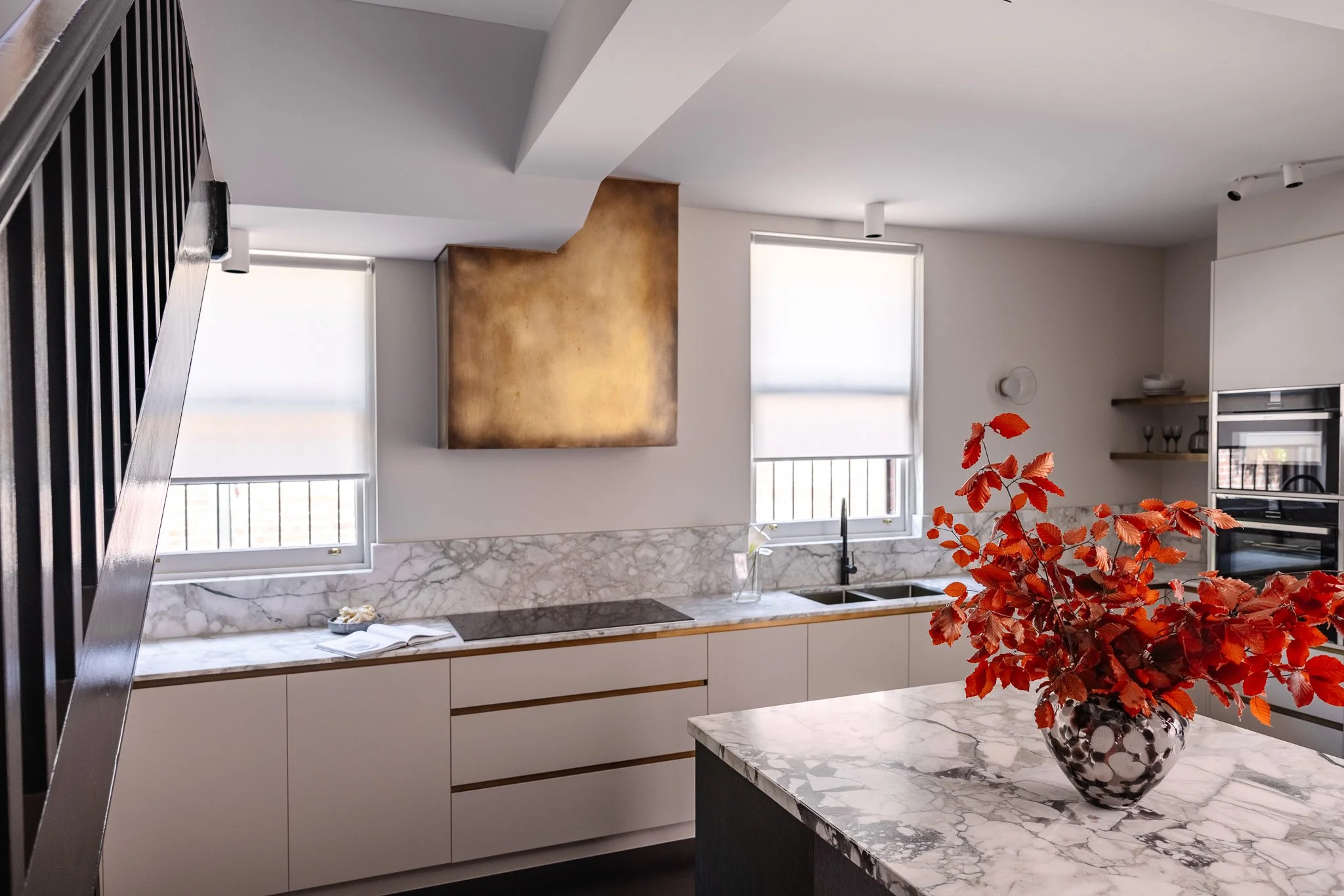 Modern kitchen with white cabinets, marble countertops, and two large windows with shades. A vase with red leaves is on a marble island.