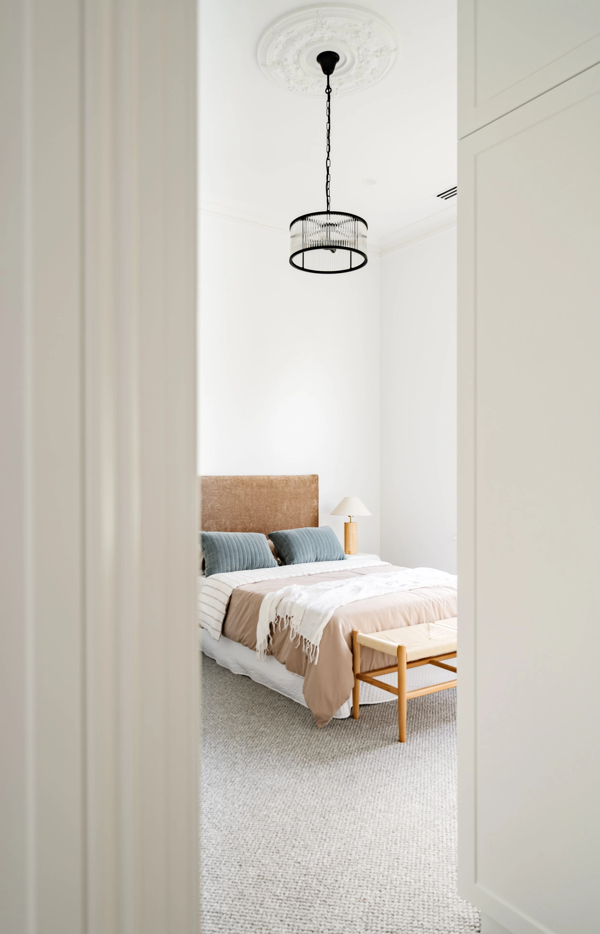Modern bedroom with a bed, blue pillows, beige and white bedding, a wooden bench at the foot, a bedside lamp, and a ceiling light fixture, seen through a doorway.