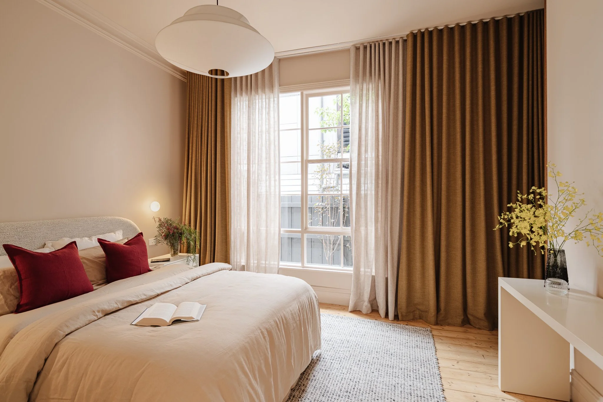 A bedroom with beige walls, a bed with cream bedding and two red pillows, and large windows with sheer and brown curtains.