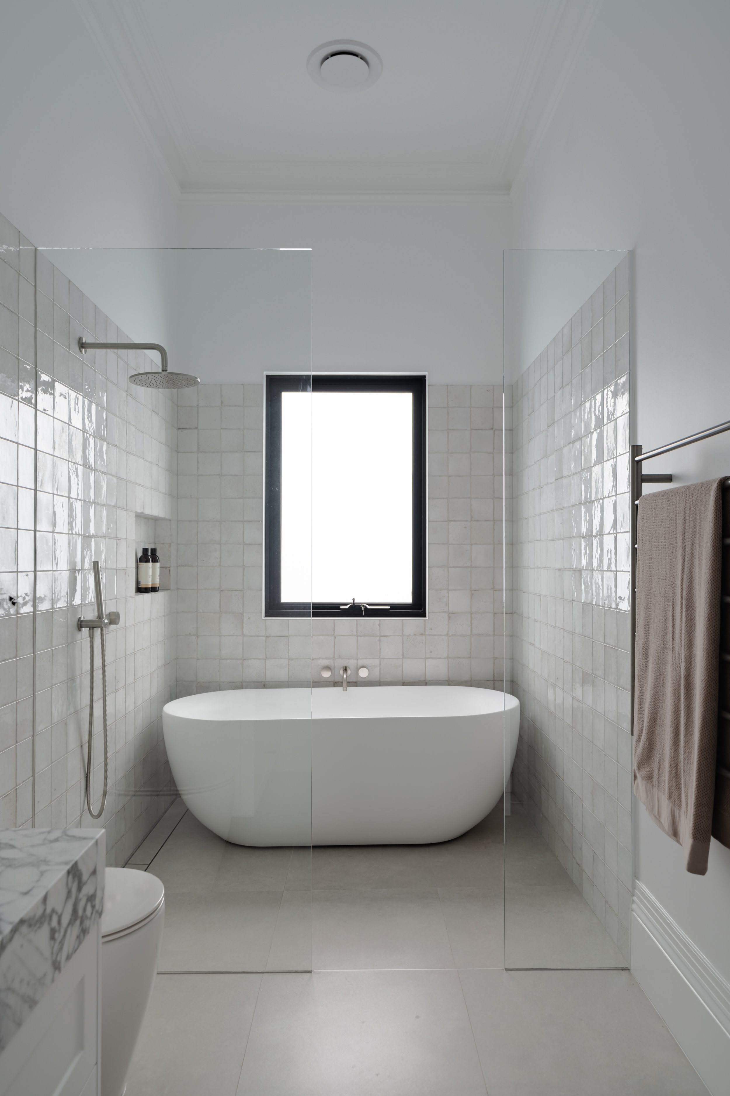 Modern bathroom with a white bathtub, a window, a rain shower, a towel rack, and a toilet, separated by glass panels.