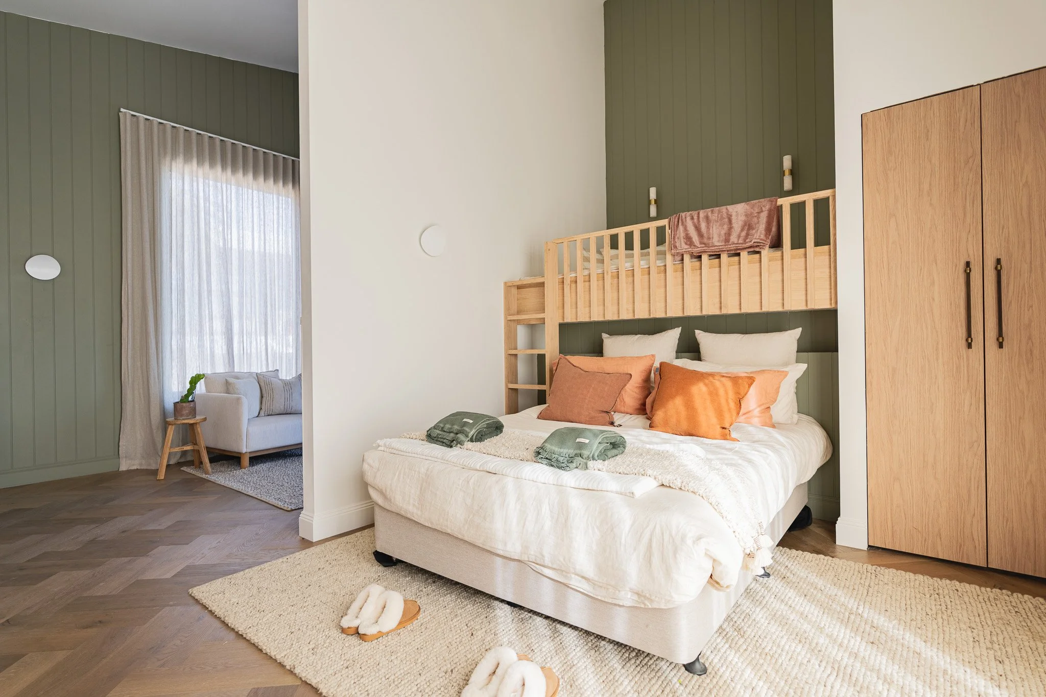 Bedroom with a bed featuring a bunk bed style headboard, beige bedding, and an assortment of orange and beige pillows. There is a beige rug on the wood floor, with slippers placed on it. In the background, a sitting area with a white couch and a smal