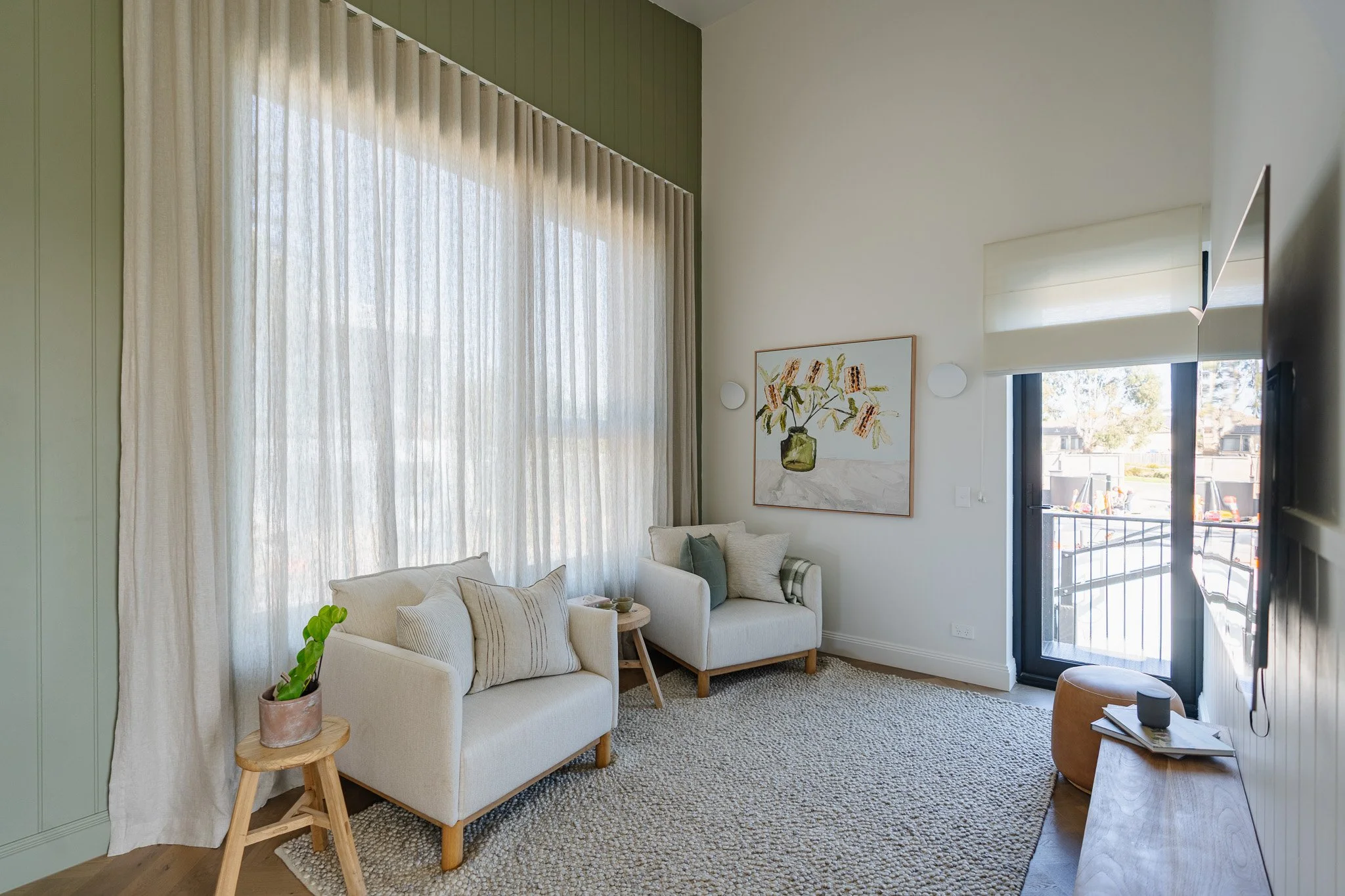 Living room with white sofa, beige armchair, wooden side table, potted plant, large window with sheer curtains, artwork with flowers, and sliding glass door leading to outside balcony.