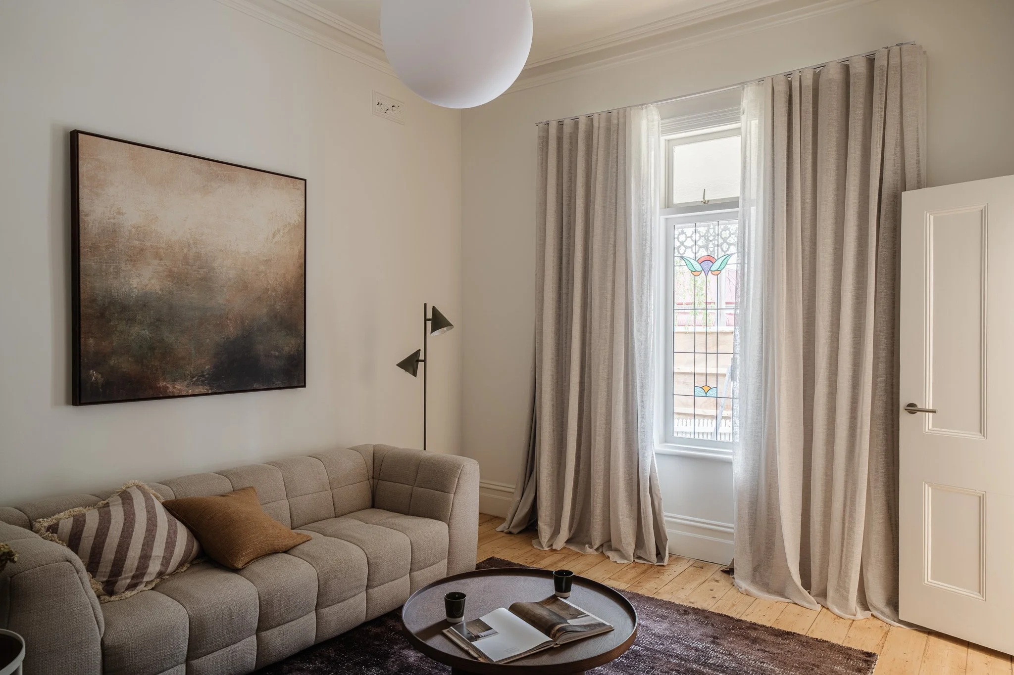 Living room with a beige tufted sofa, a round wooden coffee table with a magazine and two black cups, beige curtains, a window with stained glass, a wall-mounted lamp, a large abstract painting, and light wood flooring.