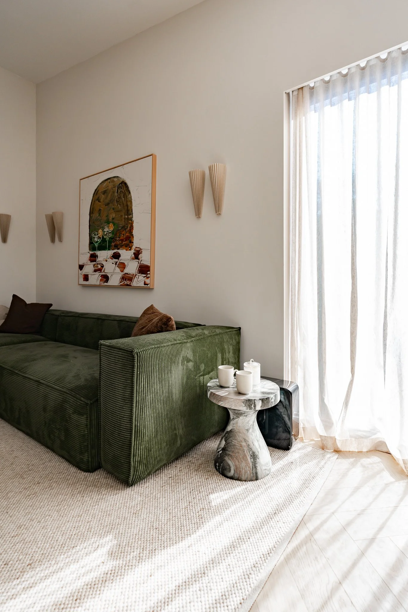 Living room corner with a green velvet sofa, a marble side table with white cups, a beige rug, and sheer curtains allowing sunlight in.