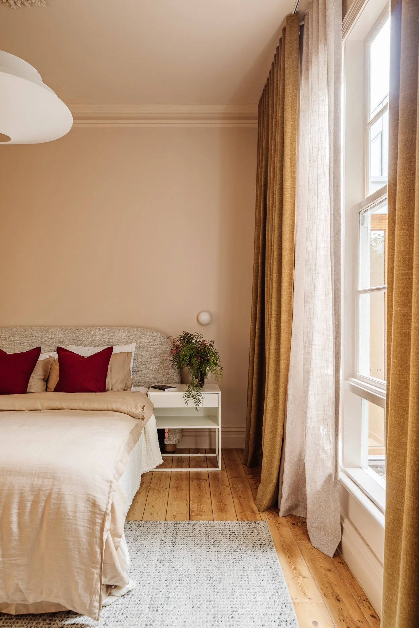 A cozy bedroom corner with beige walls, a bed with white and beige linens, red and beige pillows, a small white bedside table with a vase of flowers, and large windows with gold curtains letting in natural light.