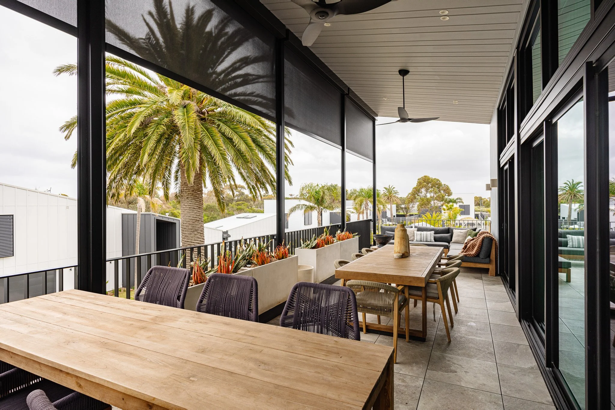 Outdoor covered balcony with wooden dining table and chairs, a seating area with sofa, potted plants with succulents, and trees including palm trees visible over the railing.