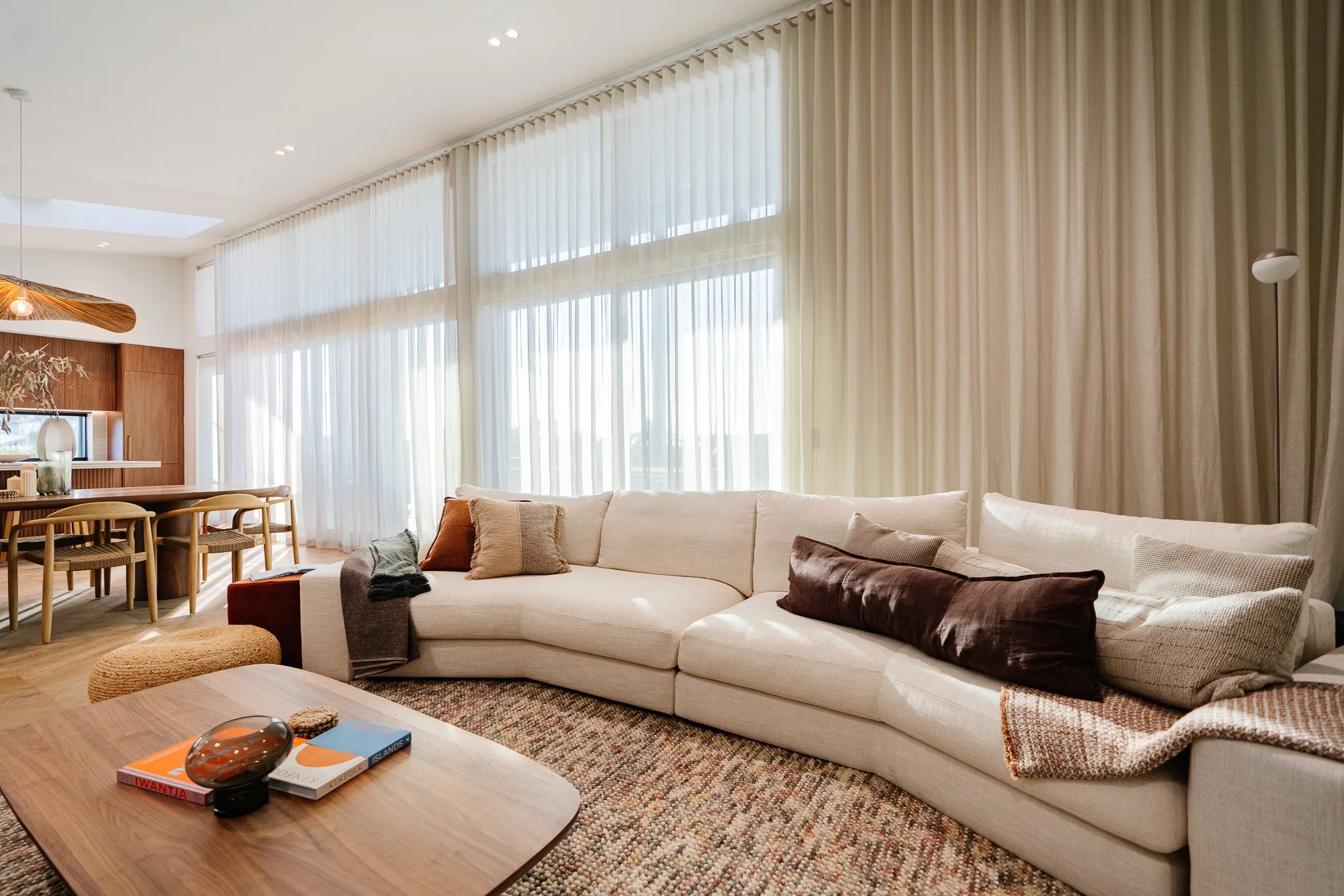 Bright living room with large cream sectional sofa, assorted throw pillows, a wooden coffee table with books and decorative items, sheer curtains, and a dining area with wooden chairs in the background.