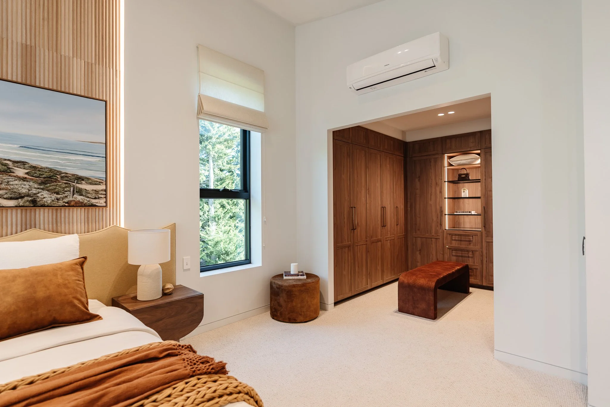Modern bedroom with white walls, a window showing green trees outside, a beige bed with brown pillows and a knitted blanket, a wooden nightstand with a white lamp, a built-in wooden closet, and a small brown sitting bench.