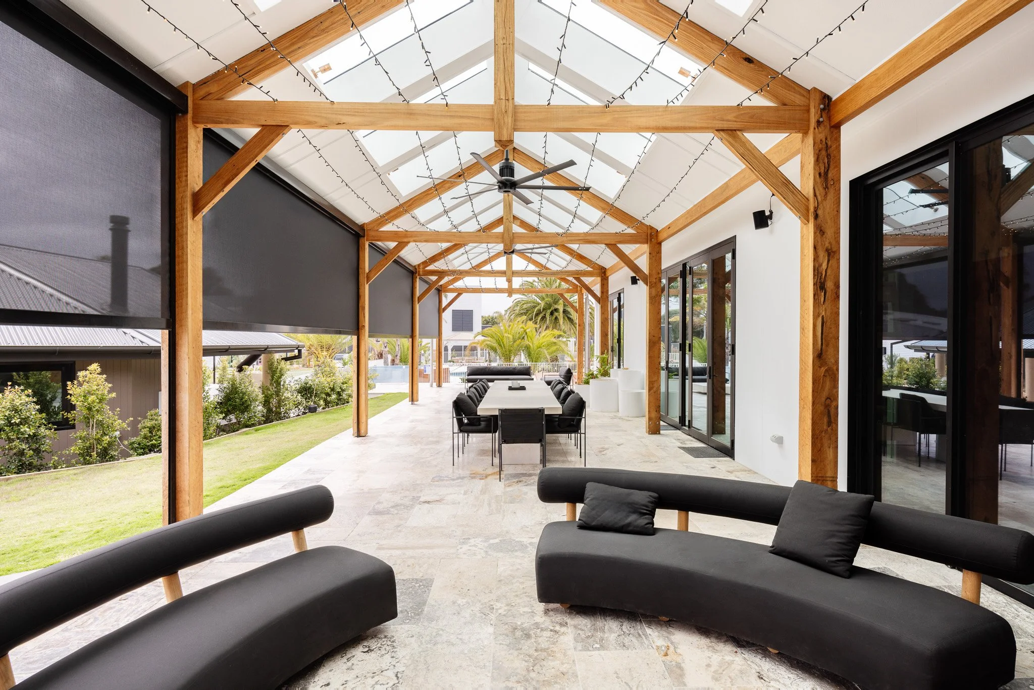 Enclosed patio with black lounge chairs, a long dining table with black chairs, ceiling fan, hanging string lights, and sliding glass doors, with a view of greenery and palm trees outside.