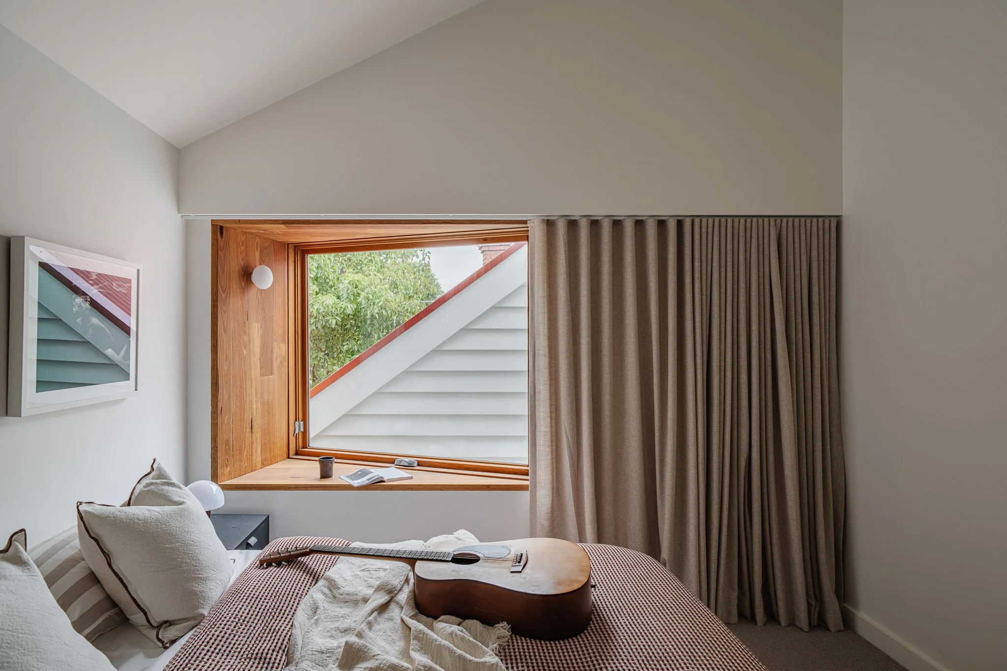 A cozy bedroom with a bed, pillows, an acoustic guitar, a window with a view of stairs and greenery outside, and beige curtains.