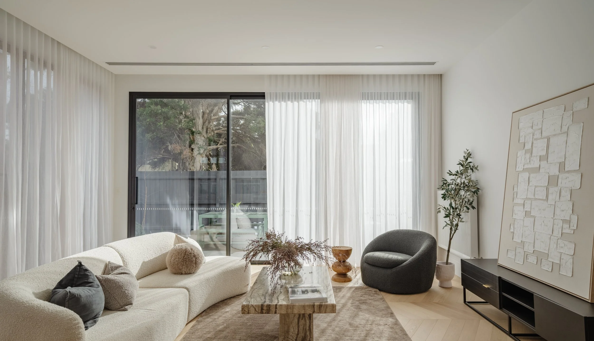 Modern living room with white sofa, black armchair, marble coffee table, large windows with white curtains, potted plant, and abstract wall art.
