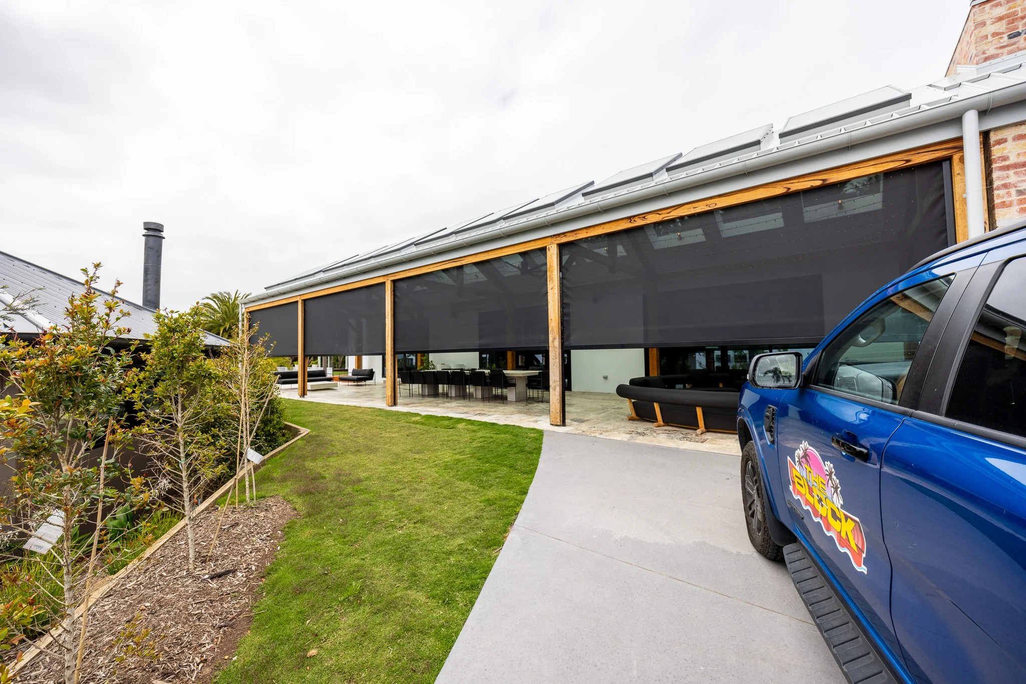 Modern building with large screened porch, outdoor seating, garden on the left, and a blue truck with a colorful logo on the right.