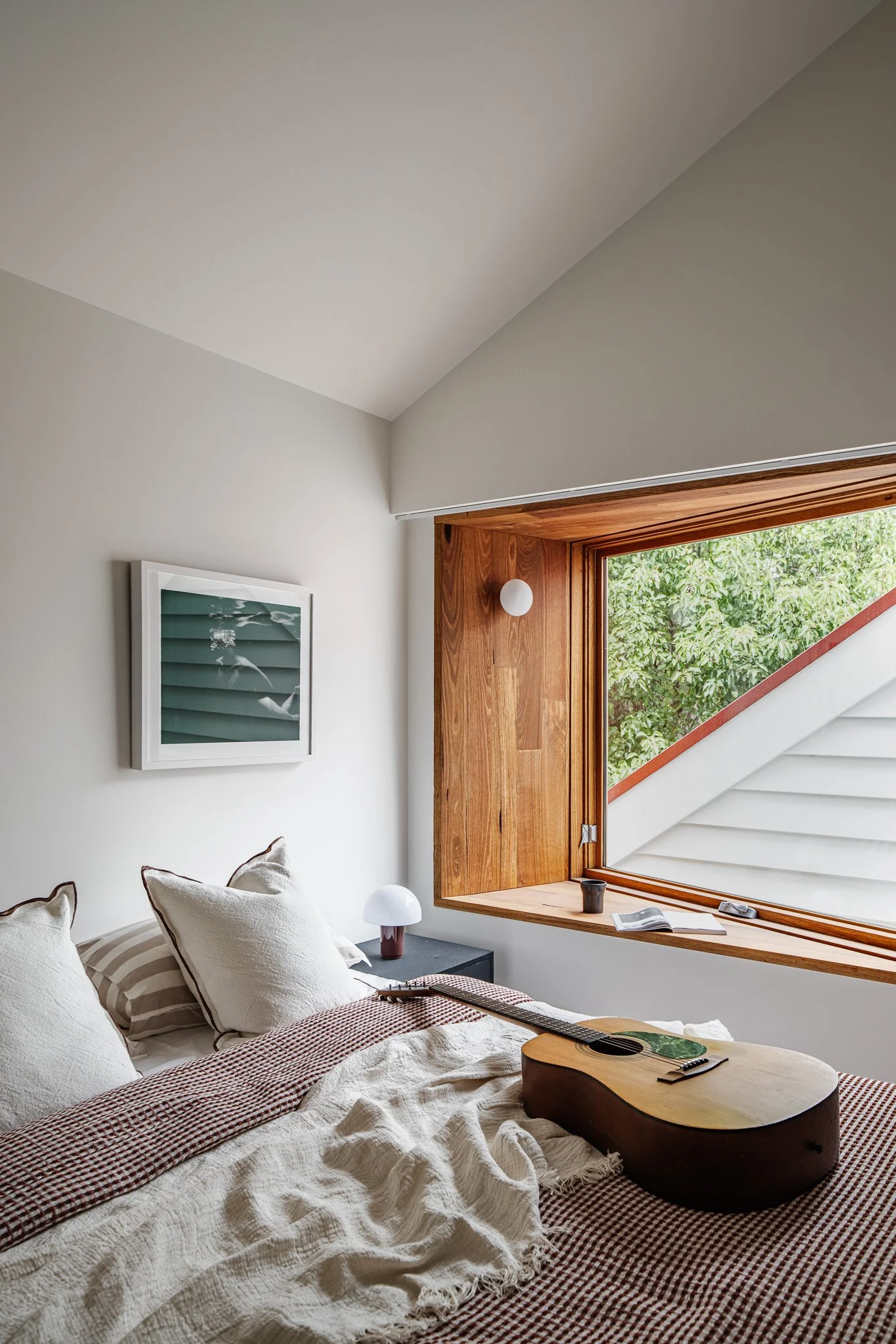 A bedroom with a bed, pillows, a guitar, a window with a wood frame, and greenery outside.