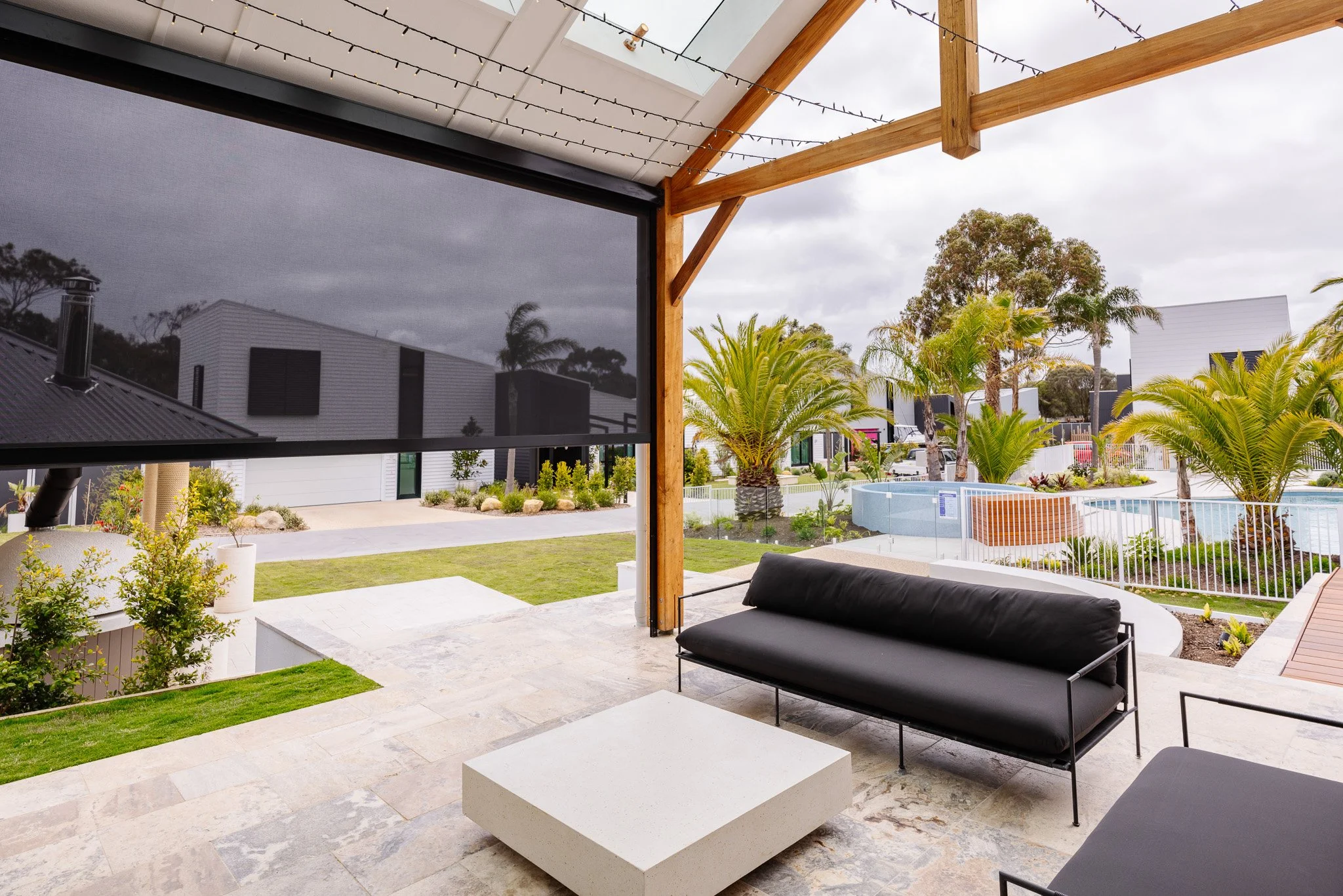 Outdoor patio with black cushioned seating and modern decor, overlooking a landscaped garden with palm trees, a small pool, and contemporary houses under cloudy sky.