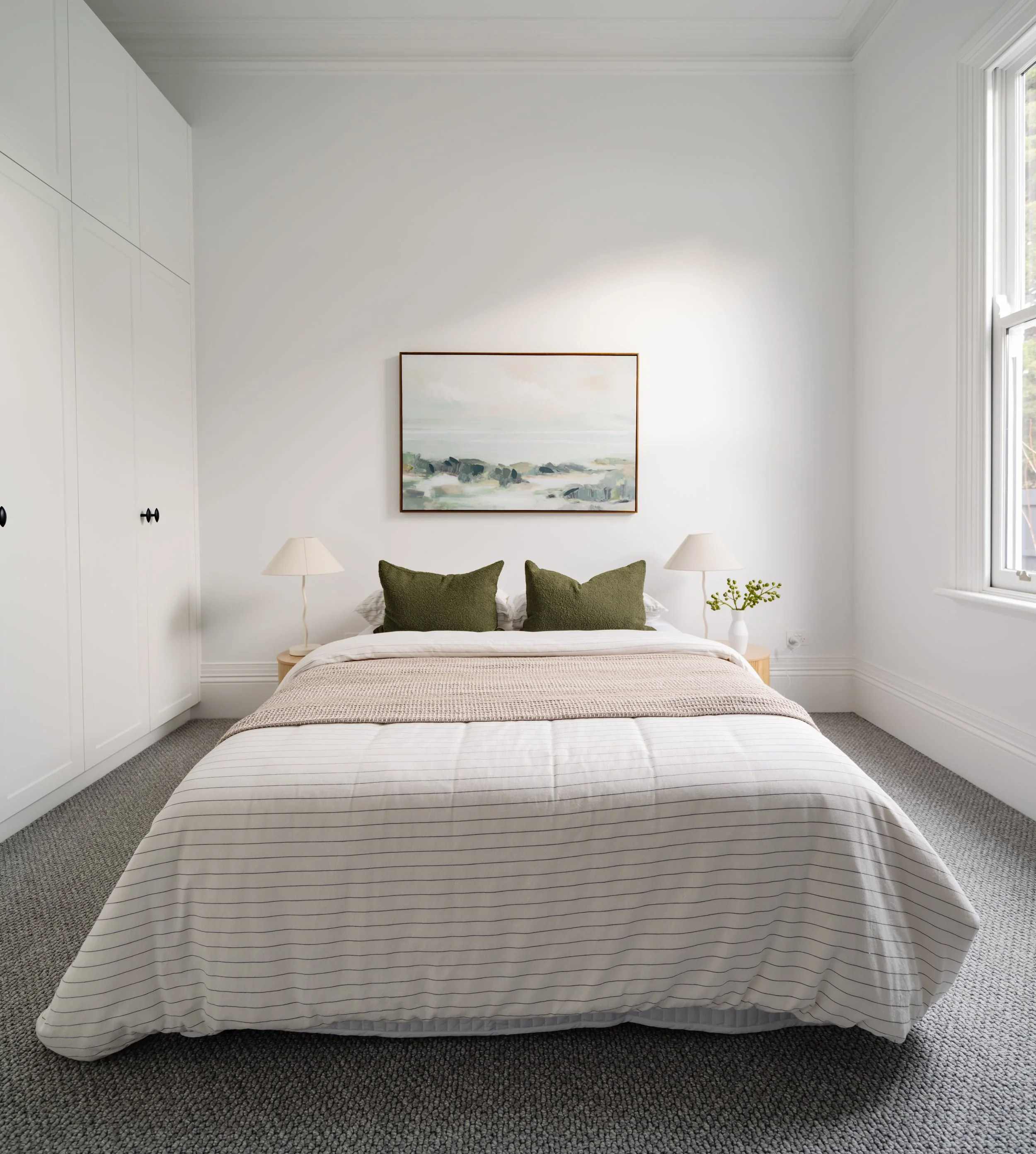 A neatly made bed with white and beige striped bedding, two green pillows, and a peach-colored blanket, in a bright bedroom with white walls, two bedside lamps, a framed landscape painting above, a window on the right, and white built-in wardrobes on