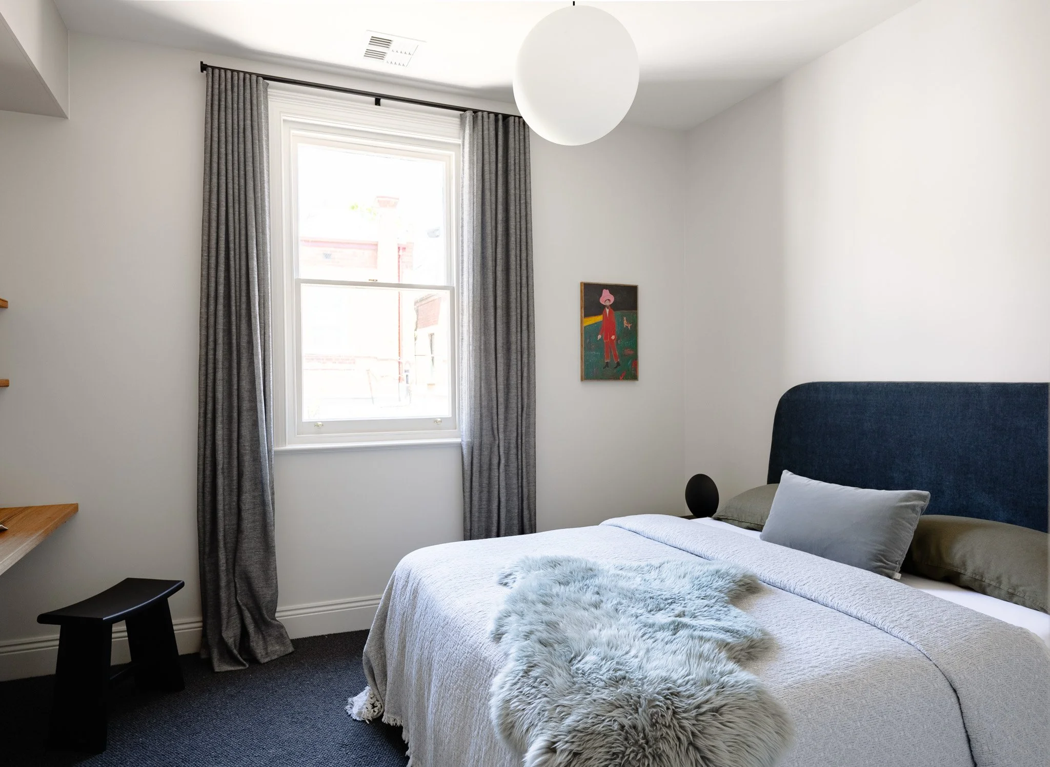 A bedroom with a bed, blue upholstered headboard, gray pillows, white blanket, furry throw, window with gray curtains, a small black stool, a floating shelf, a round white ceiling light, and a colorful painting on the wall.