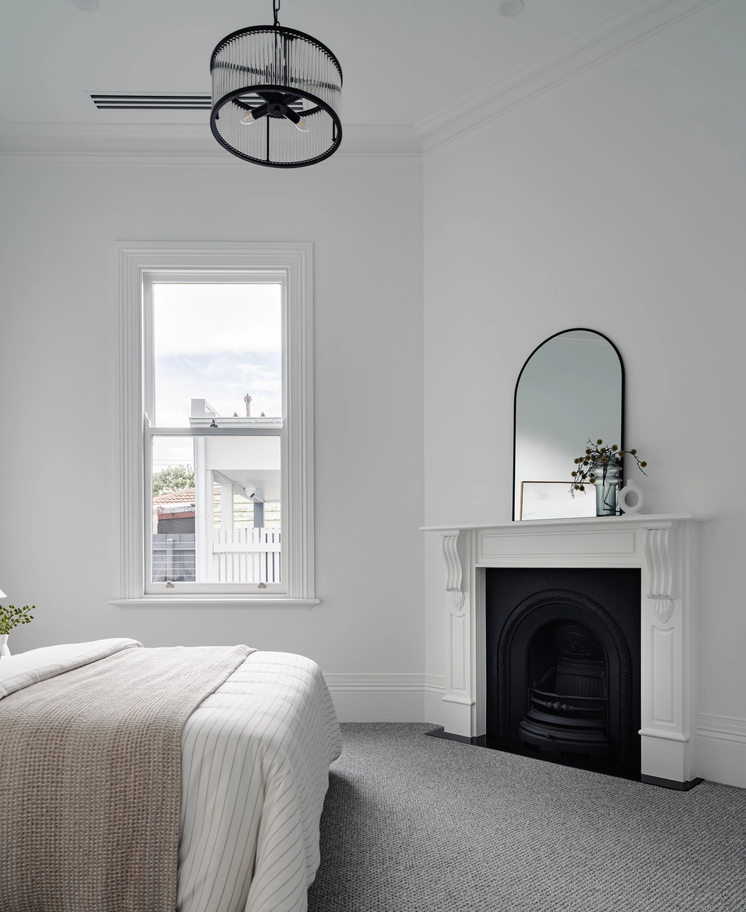 A bedroom with a large window, a white bed with a beige blanket, a white fireplace with a mirror and decorative items, and a ceiling light fixture.