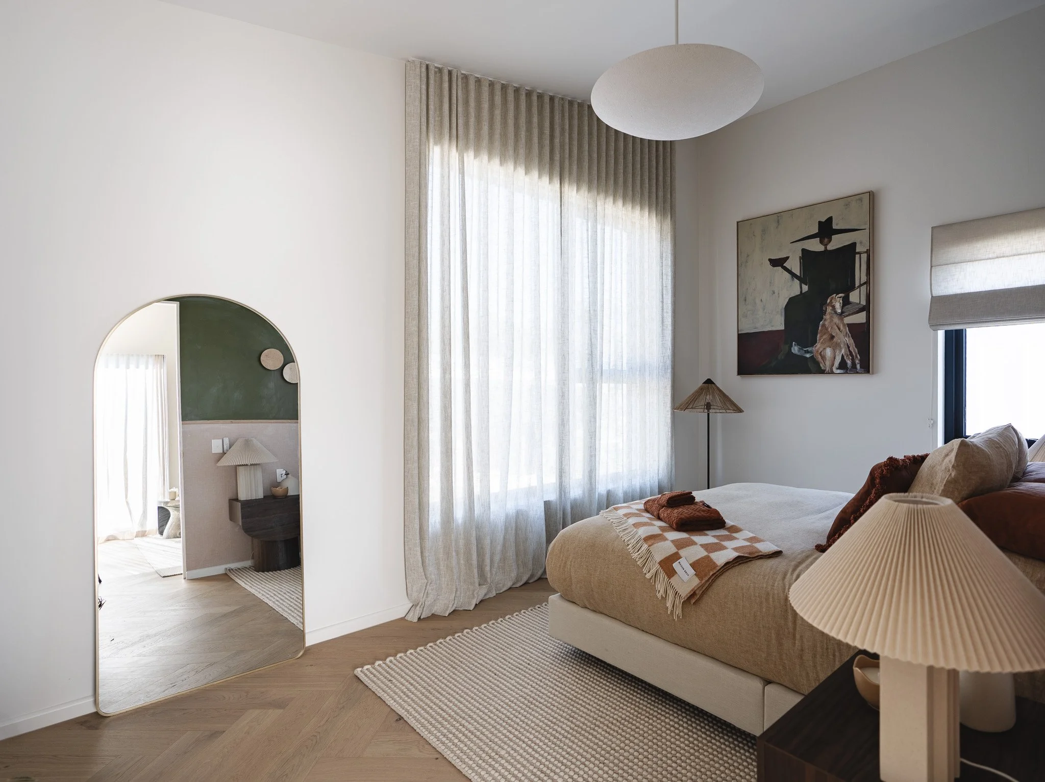 A cozy bedroom with a large bed, beige bedding, pillows, and a throw blanket. There is a wooden nightstand with a pleated lampshade, a wall-mounted mirror, a window with sheer curtains allowing sunlight, and a modern painting above the bed. The room 