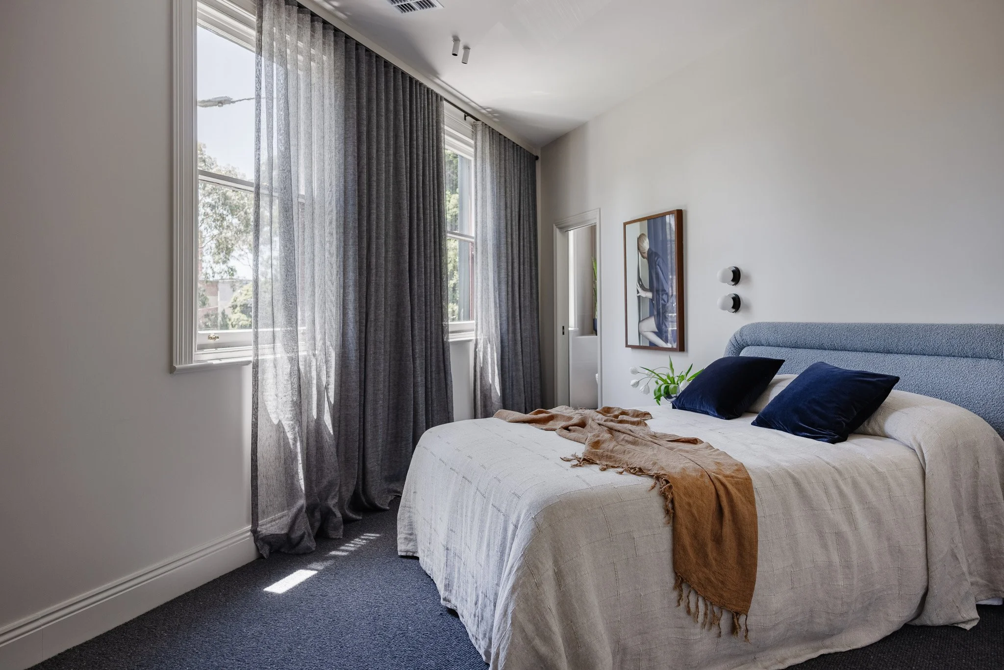 A bedroom with a bed, navy blue pillows, beige bedding, a brown throw blanket, and large windows with dark gray curtains, sunlight streaming in, and artwork on the wall.