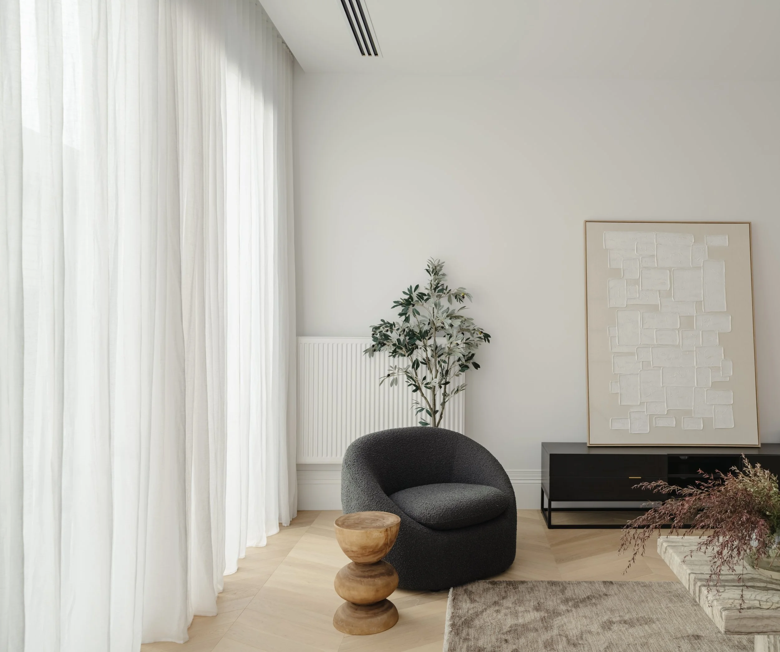 Modern living room with white curtains, a black round armchair, a small wooden side table, a tall indoor plant, a dark wood console table, abstract wall art, and a beige area rug.