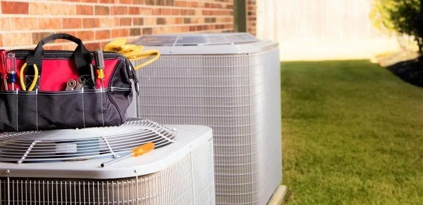 Outdoor scene showing two HVAC units next to a brick wall, with a tool bag on top of one unit and a yellow garden hose in the background near a grassy yard.