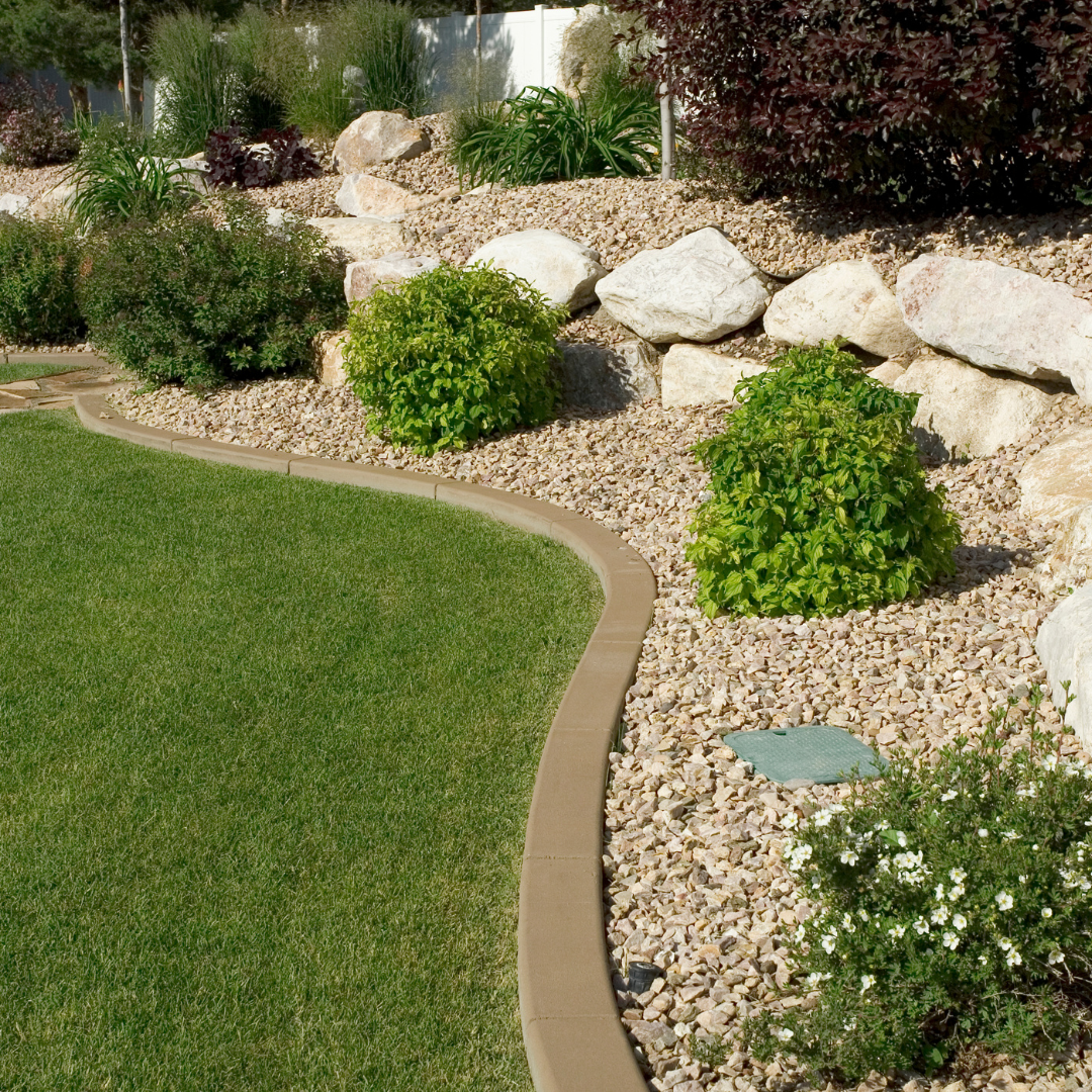 A landscaped backyard with a green lawn, decorative rocks, shrubbery, and plants along a curved border.