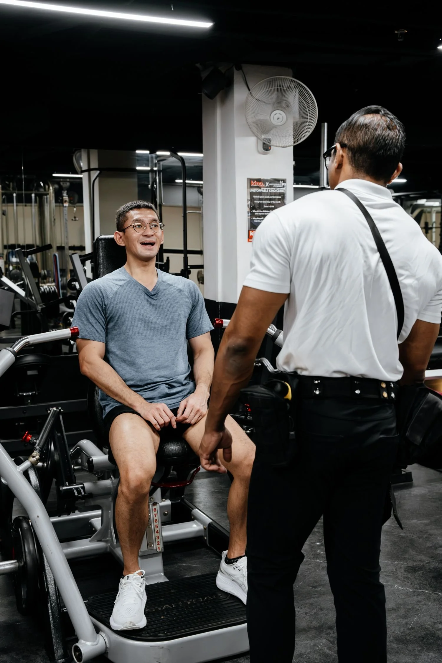 A man sitting on exercise equipment in a gym, conversing with his personal trainer