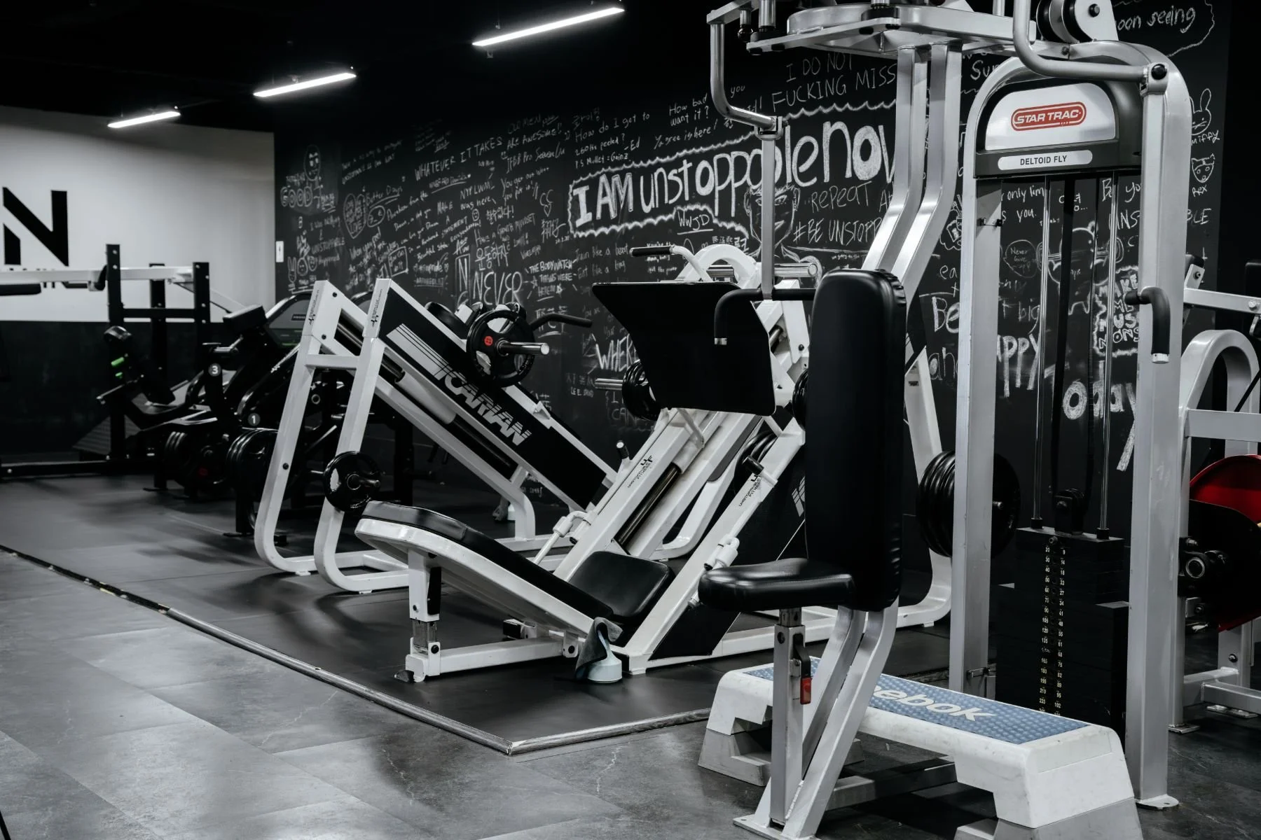 Unstoppable Ftiness. Gym located at Shenton Way, with various exercise equipment including weight machines and cardio machines, black and white color scheme, chalkboard wall with motivational messages.
