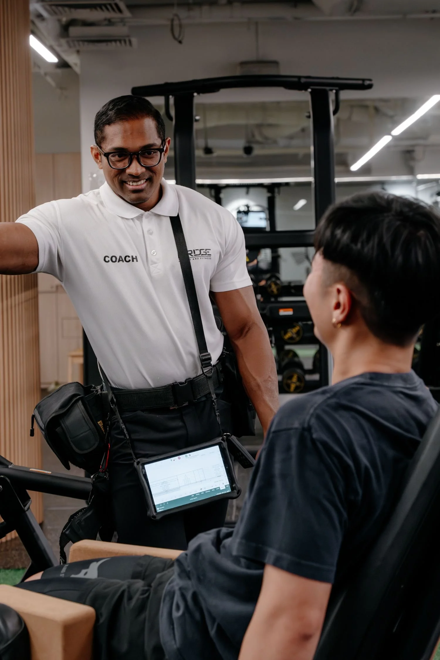 Head Coach of Bridge Health and Fitness, Khalis, with an electronic device hanging around his neck to monitor and record trainees progress. The person on the machine has short hair and is wearing a black shirt, listening attentively.